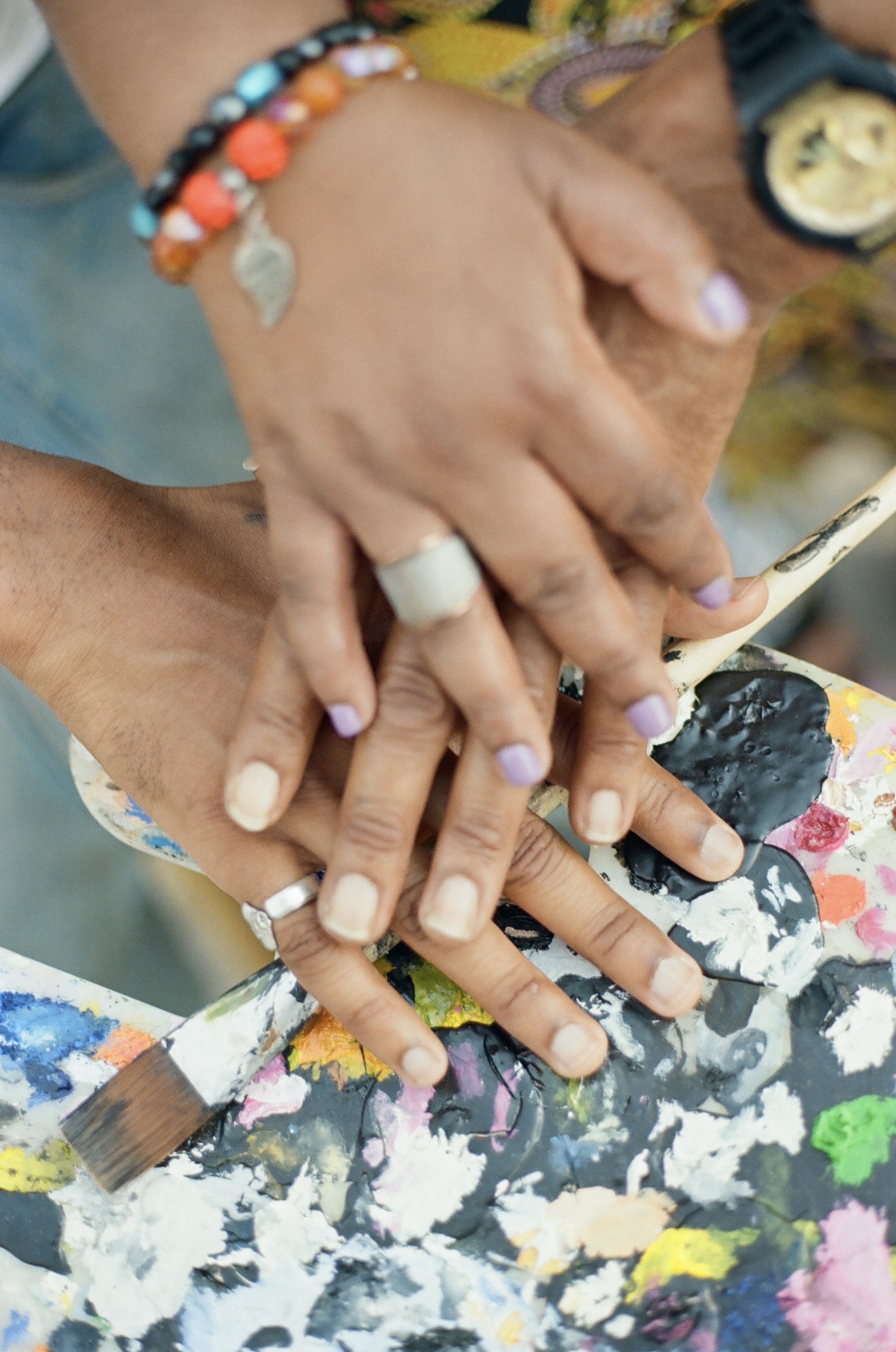 Close-up of multiple hands with painted nails and jewelry placed on a colorful, textured surface with paint splatters, and an artist's brush in the background.