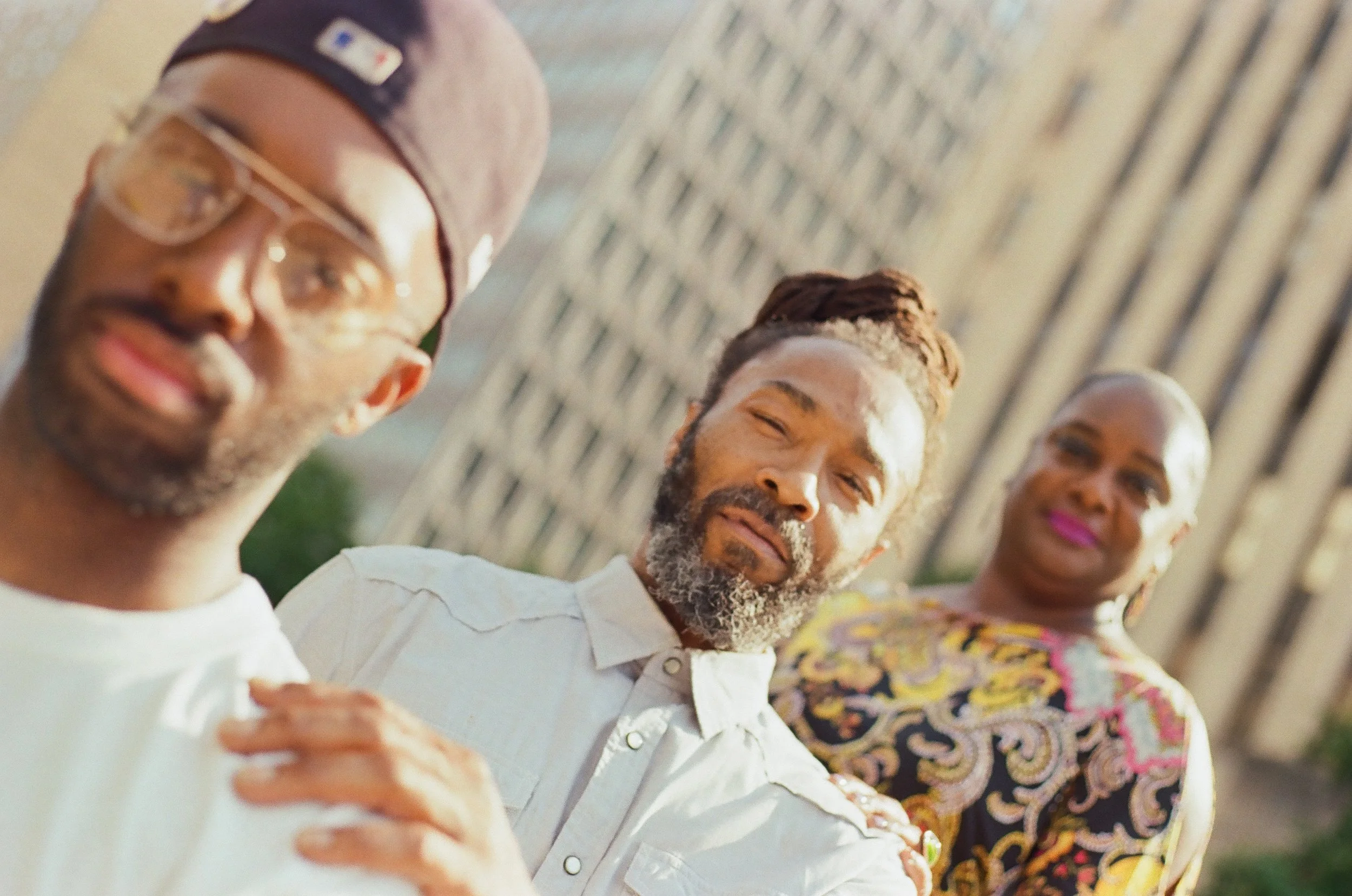 Three adults smiling and standing outdoors on a sunny day, with blurred background of a fence.