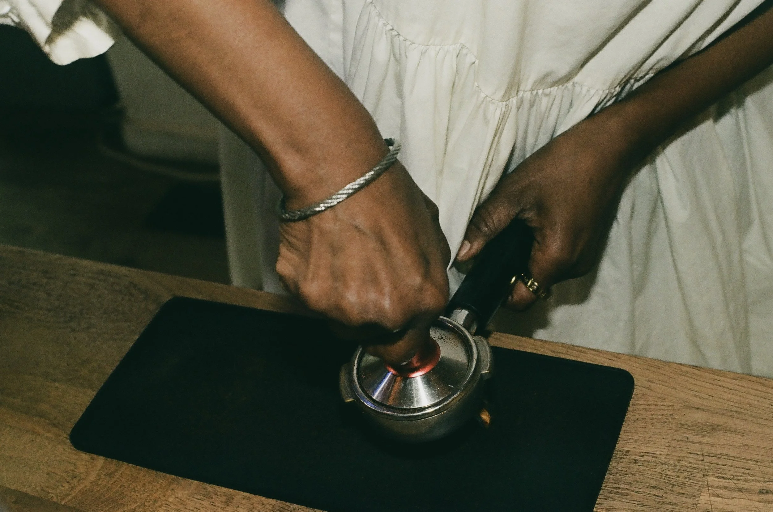 A person with dark skin is using a blowtorch to melt a metal ring on their wrist. The person is wearing a silver bangle and a white garment, and is working on a wooden surface.