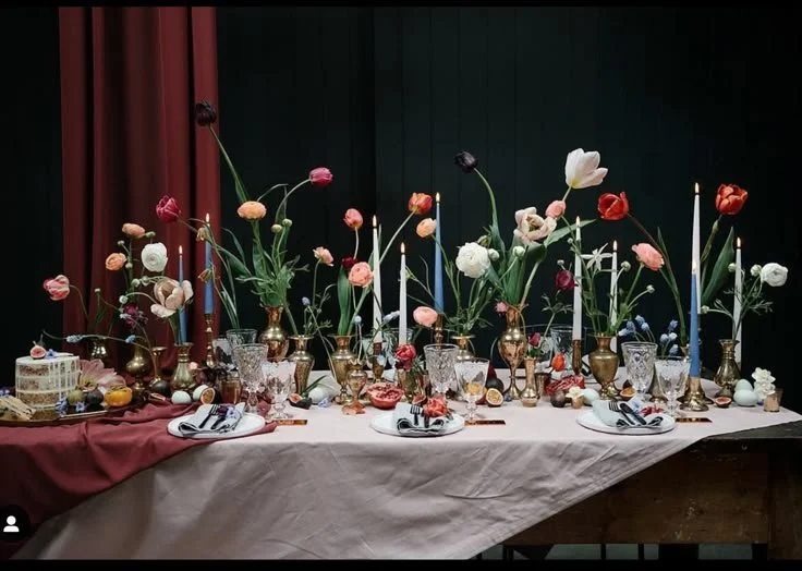 A table decorated with tall flowers in vases, candles, crystal glasses, and plates with napkins, set for a meal.
