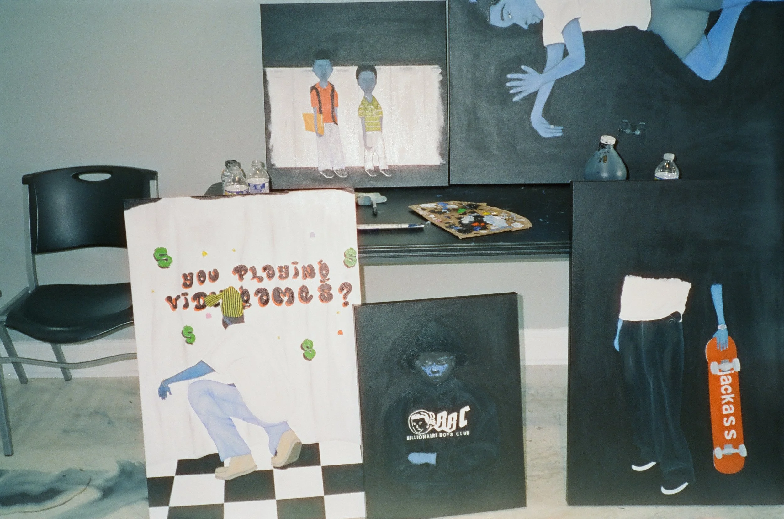 A wall with several paintings and posters including one of two kids standing with backpacks, another of a person lying on their side, and a third of a person playing basketball. There are also water bottles, a palette, brushes, and various art supplies on a table below the paintings.