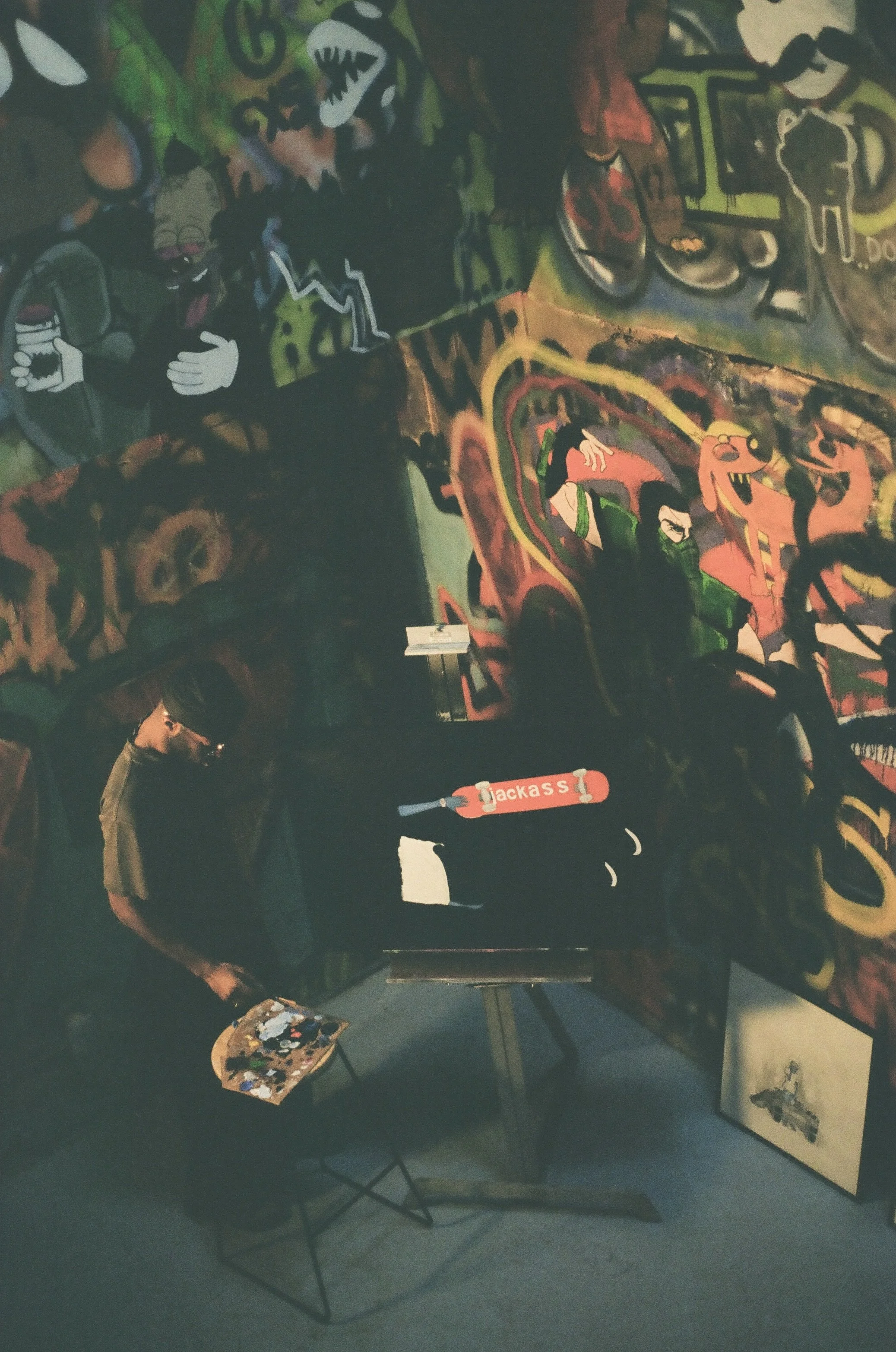 A person in dark clothing and glasses stands in front of a colorful graffiti wall, holding a paint palette, possibly an artist. The graffiti features various vibrant characters and abstract designs, including a cartoon dragon and several others. There is a small easel or stand with a paint palette on it, set on a dark floor.