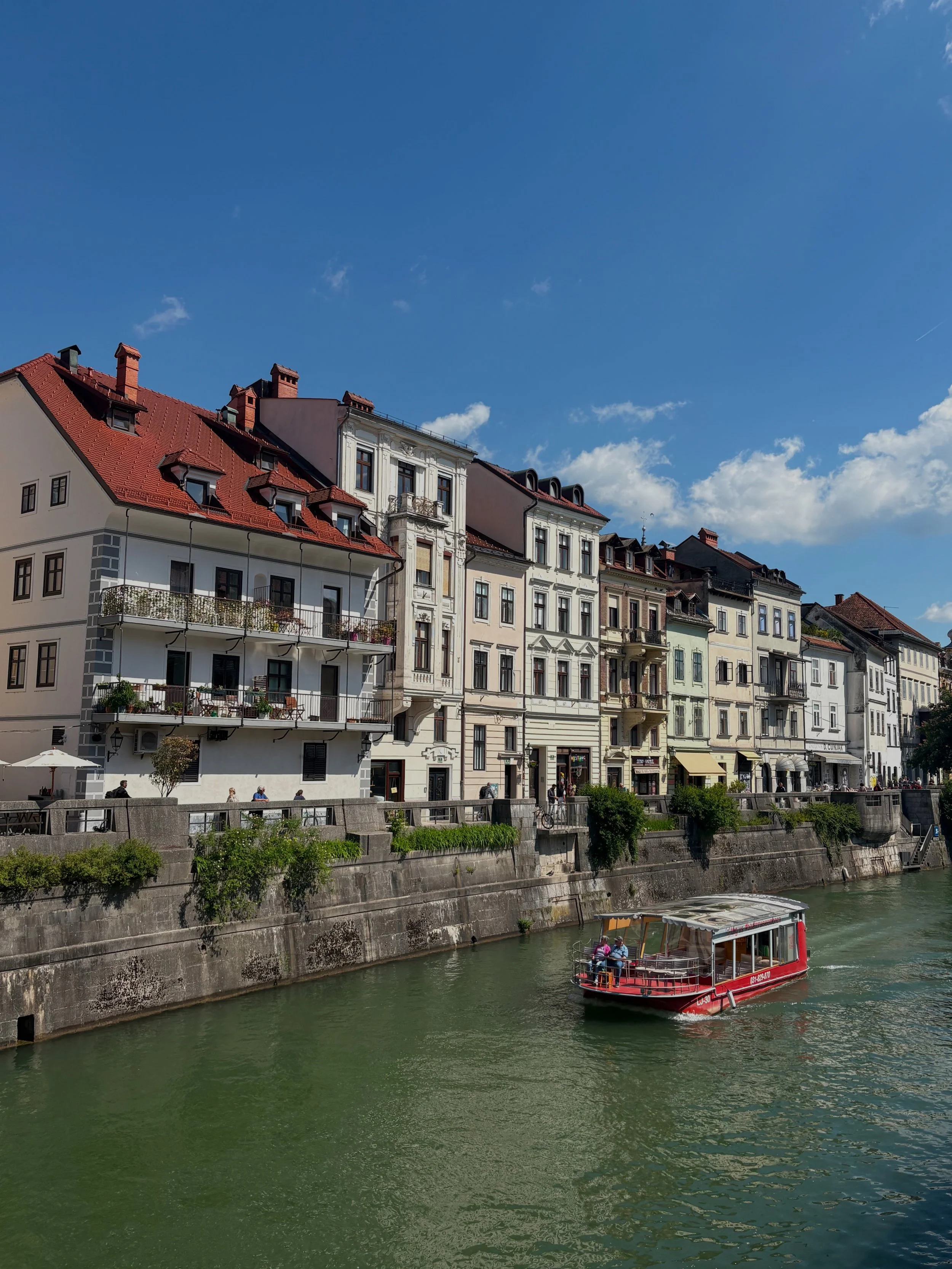 Red tourist boat cruising the Ljubljanica River past pastel townhouses in Ljubljana's Old Town, Slovenia