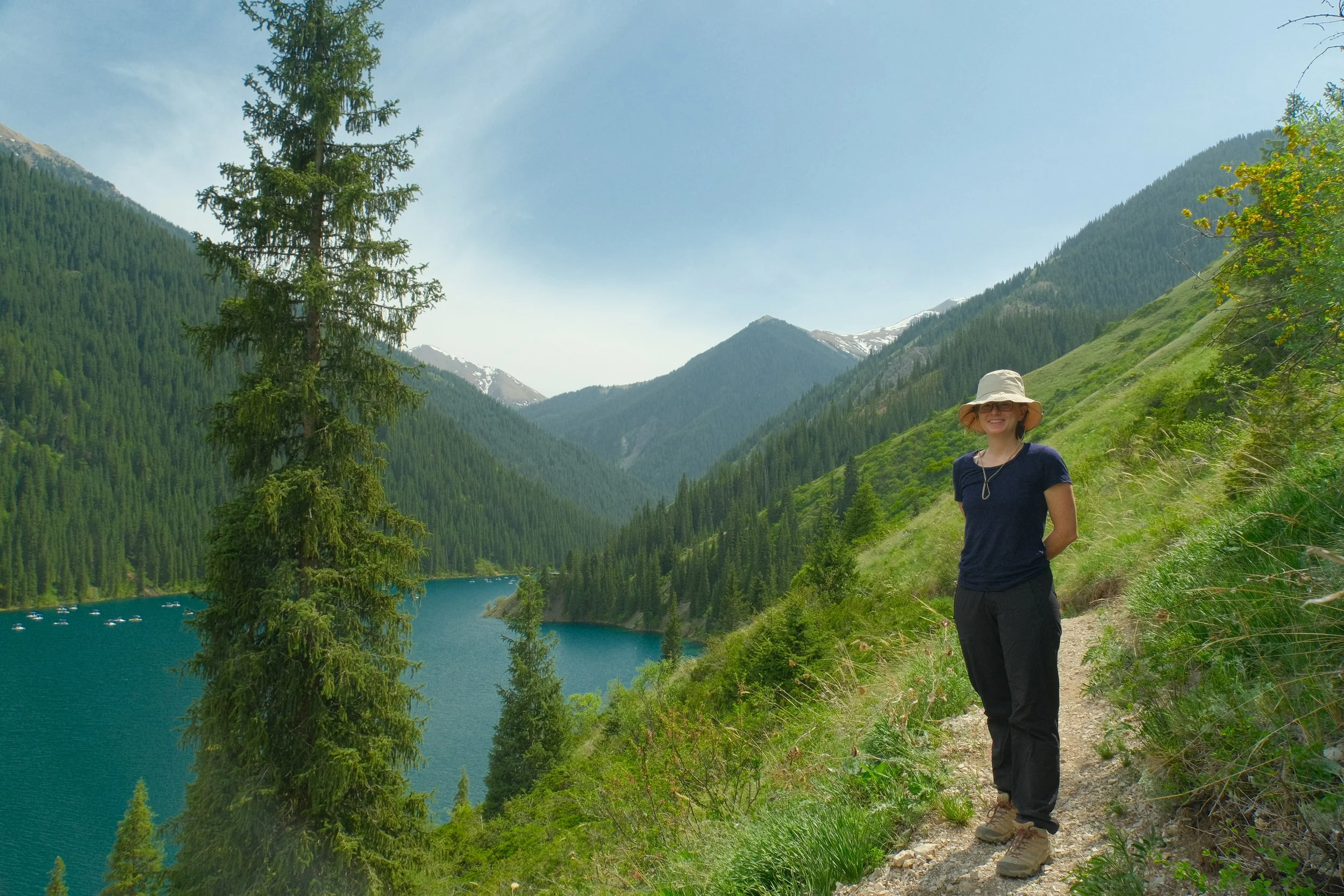 Traveler on a hiking trail above the turquoise waters of Kolsai Lake surrounded by pine forest and snow-capped mountains, Kazakhstan