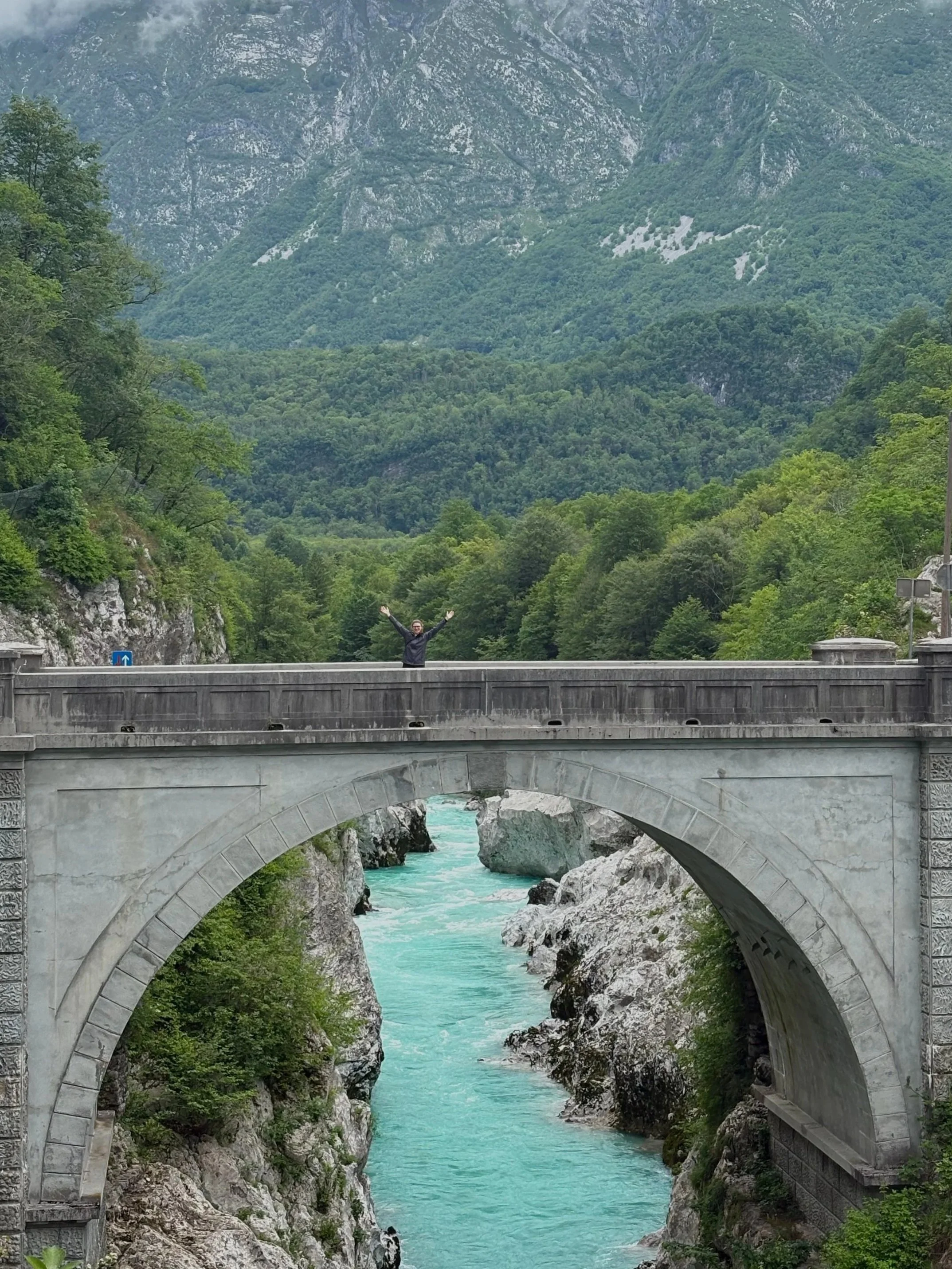 Person standing on Napoleon's Bridge with arms raised, overlooking the turquoise Soča River gorge and Julian Alps near Kobarid, Slovenia
