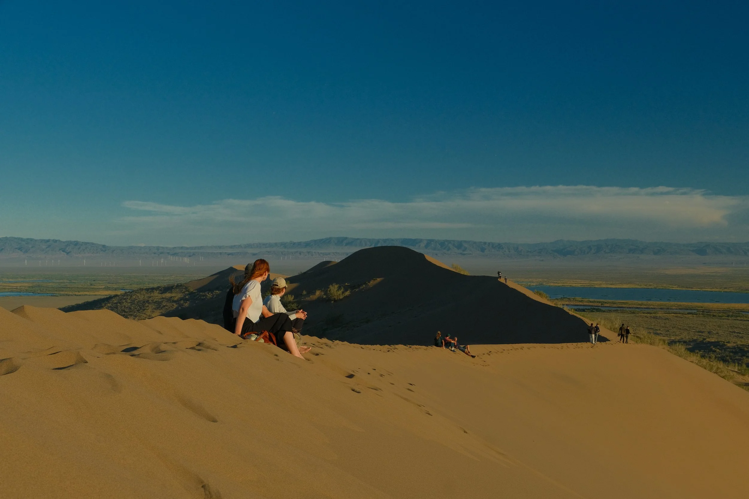 Two travelers sitting on the Singing Dunes at golden hour overlooking the steppe and mountains of Altyn-Emel National Park, Kazakhstan