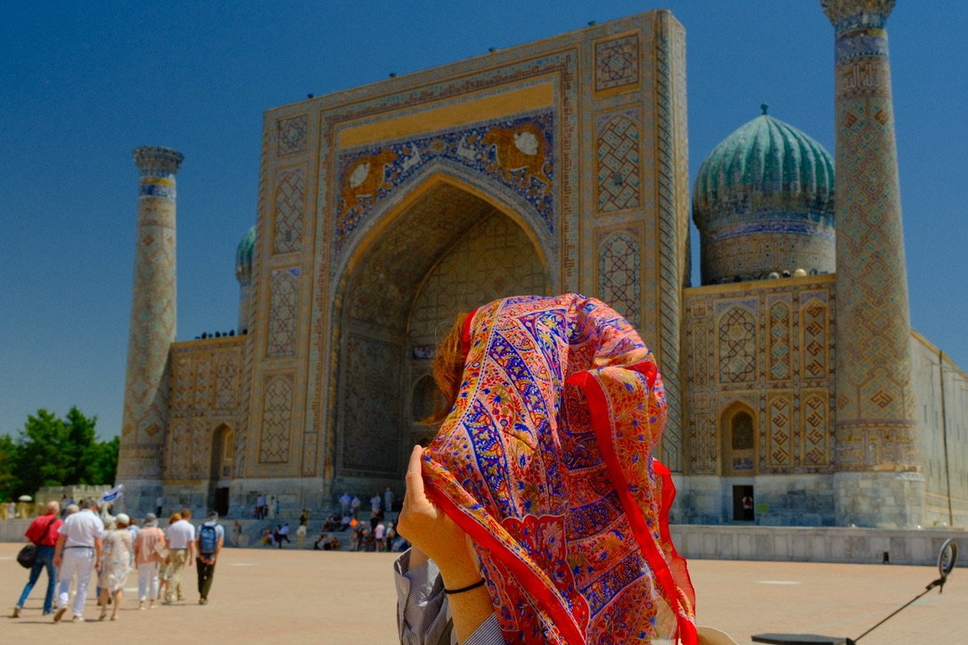 Woman holding a colorful patterned headscarf in front of Sher-Dor Madrassa at Registan Square, Samarkand, Uzbekistan