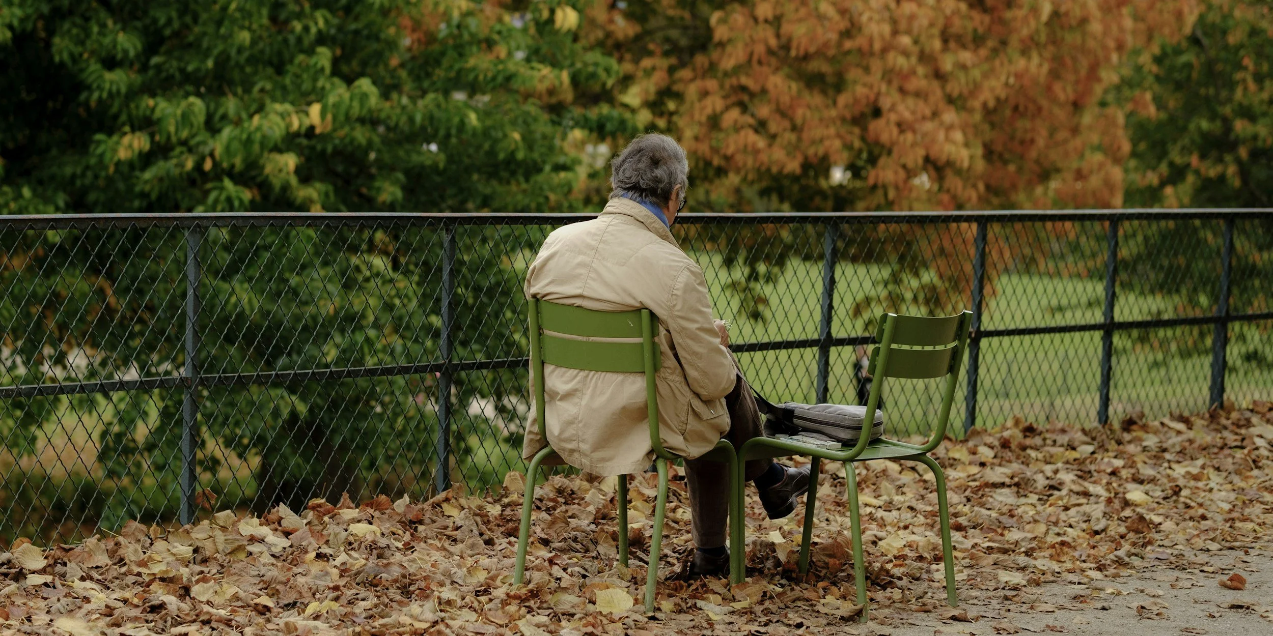Older man sitting on a green metal chair on a leaf-covered ground, facing away, beside another empty chair, with autumn trees in the background.