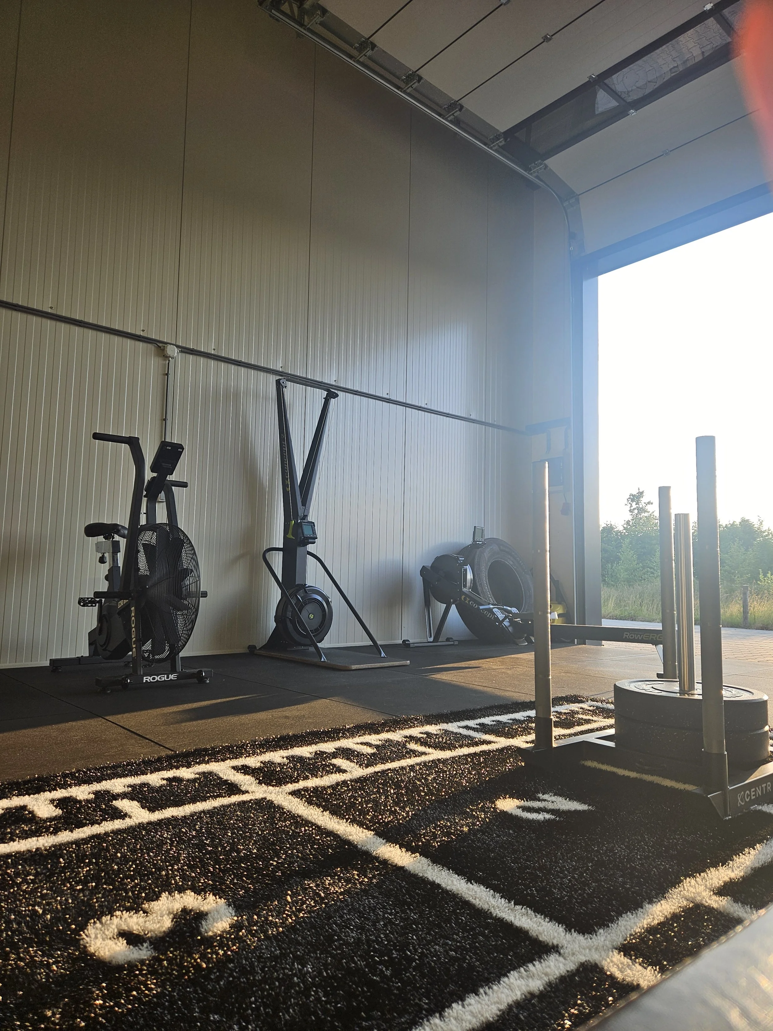 Fitnessruimte met cardioapparatuur en een houten vloer met sportieve markeringen, open deur naar buiten met groene bomen op de achtergrond.