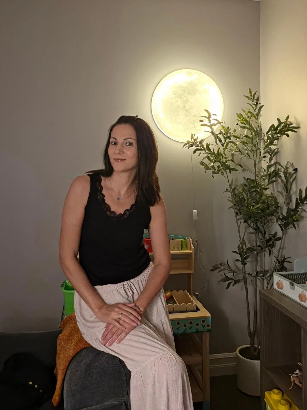 A woman with shoulder-length dark hair wearing a black sleeveless top sitting on a gray ottoman in a room with a large circular moon lamp on the wall and a tall potted plant to her right. There is a small wooden table with children's toys behind her.