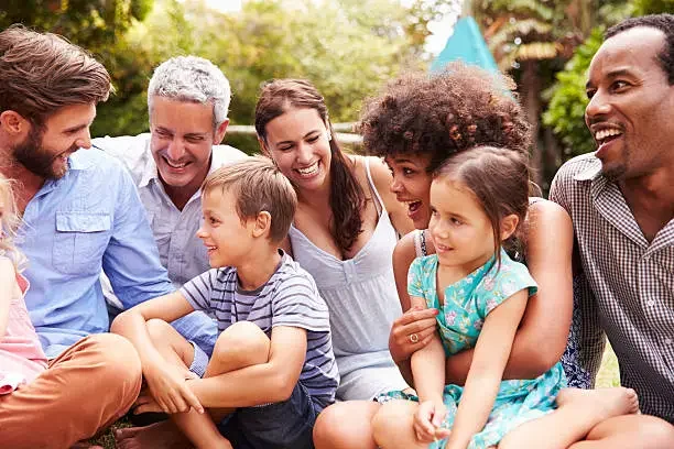 A diverse group of children and adults sitting outdoors on grass, smiling and enjoying each other's company during daytime.