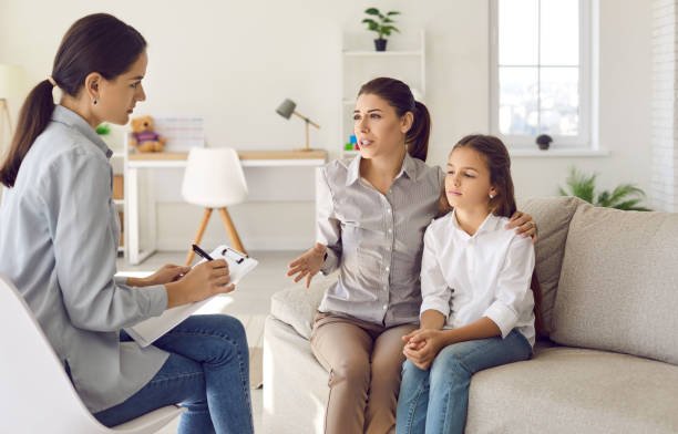 A woman and a girl sitting on a beige sofa having a discussion with a therapist in a bright, modern room.