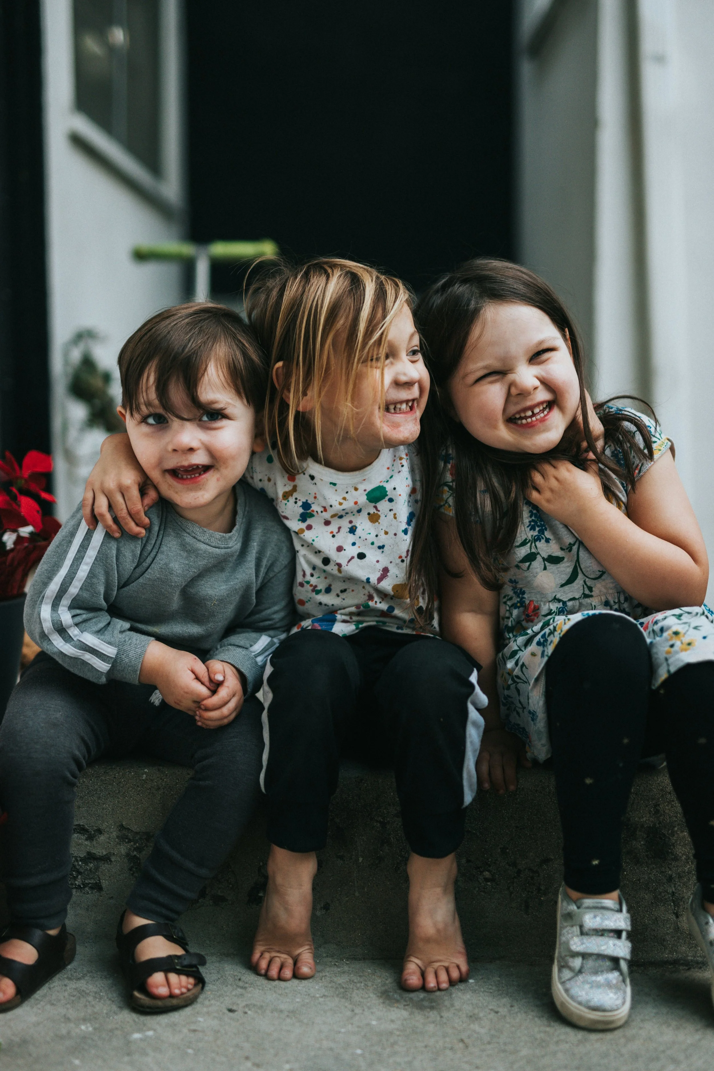 Four children sitting on the ground, smiling and hugging each other, with their shoes visible. They are dressed casually and appear happy.
