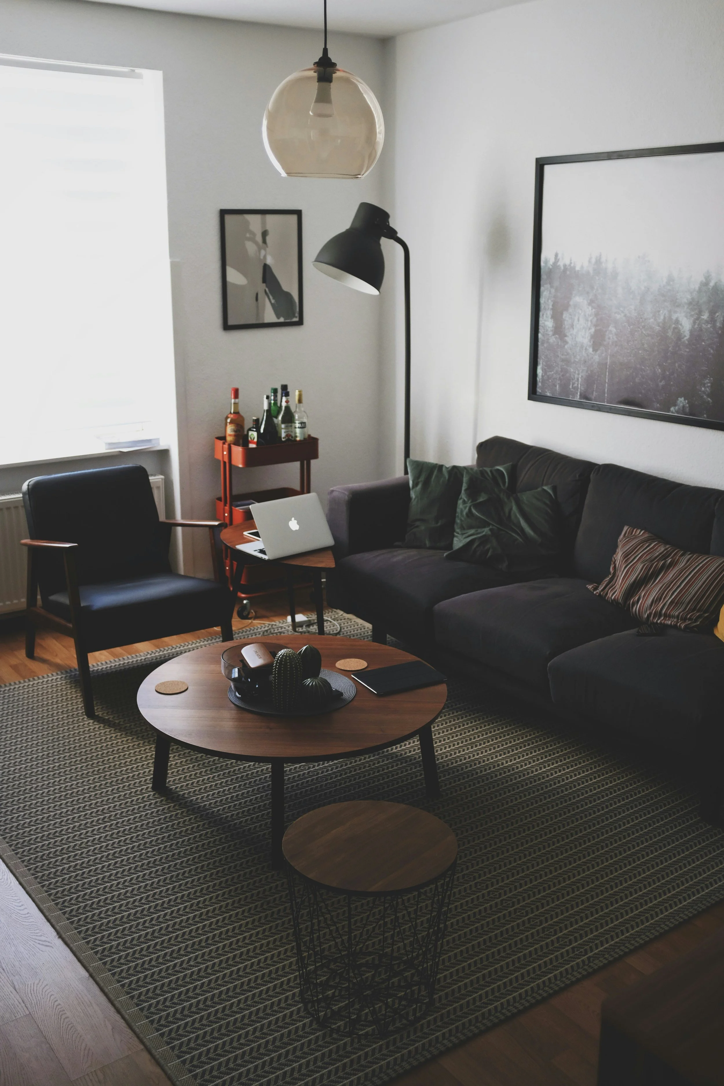 A living room with a black sofa, pillows, a round wooden coffee table with decor items, a patterned rug, a black armchair, a laptop, a side table with bottles, wall art, large window, floor lamp, and ceiling light.