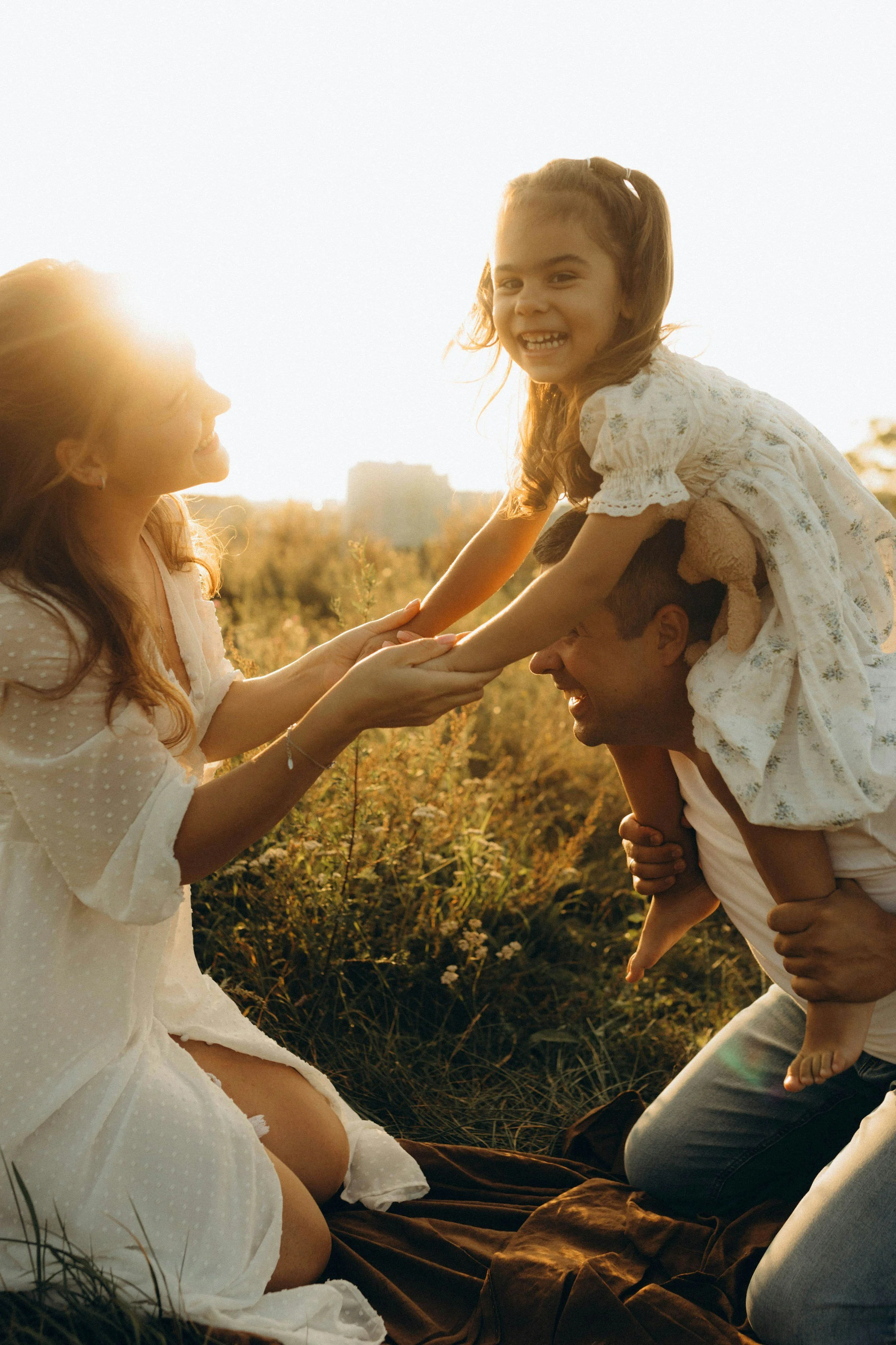 A family playing outdoors during sunset. A woman and a man are holding a young girl on the man's shoulders. The girl is smiling and reaching out to the woman, who is smiling back. They are surrounded by tall grass and wildflowers, with a cityscape in the distance.