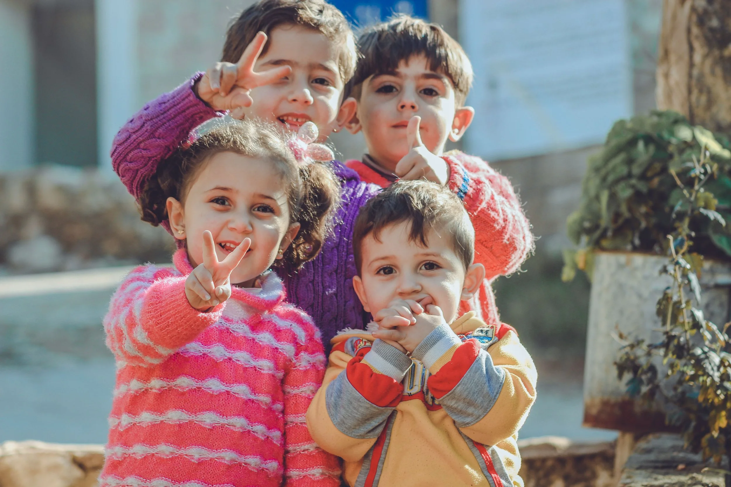 Five children outdoors posing for a photo, smiling and making peace signs. They are dressed in colorful sweaters, with a tree and a fence in the background.