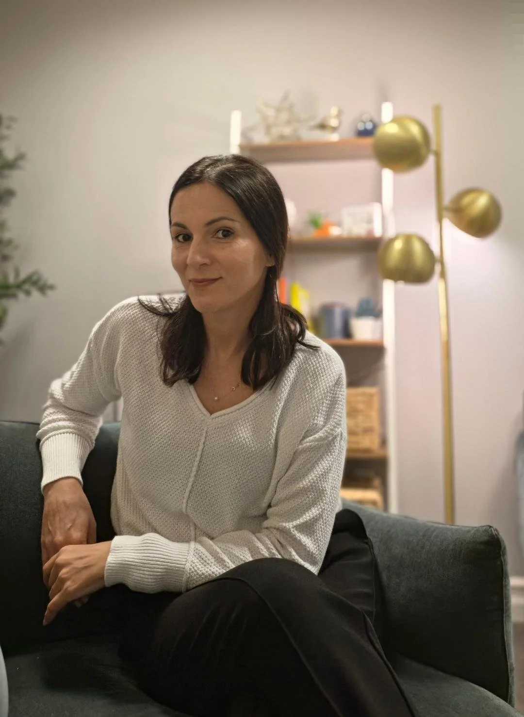 A woman with shoulder-length dark hair, wearing a white sweater, sitting on a dark green couch in a cozy living room with a bookshelf and a gold floor lamp in the background.