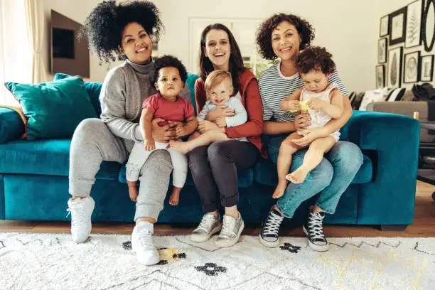 Five women and three young children sitting on a teal couch in a living room, smiling at the camera.