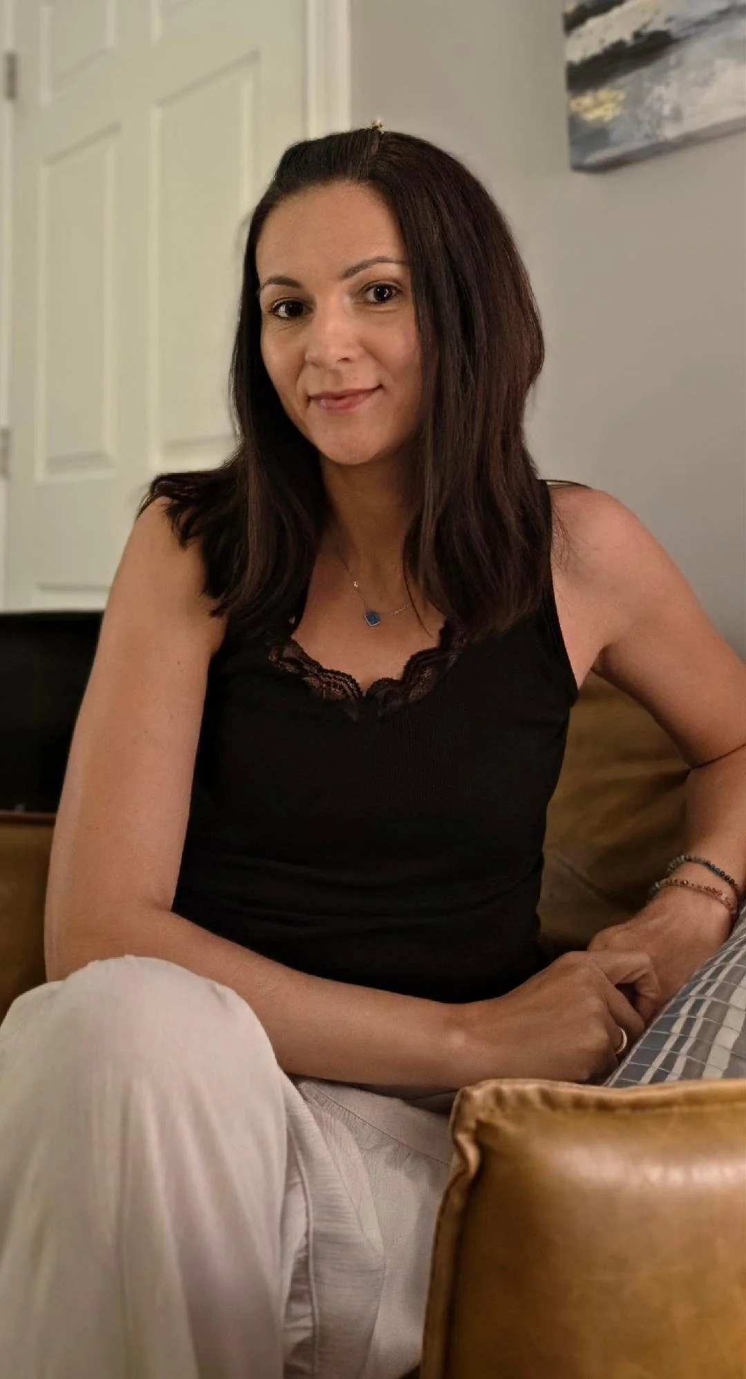 A woman with shoulder-length dark brown hair, wearing a black top with lace at the neckline, sitting on a couch with her right arm resting on the armrest, smiling at the camera.