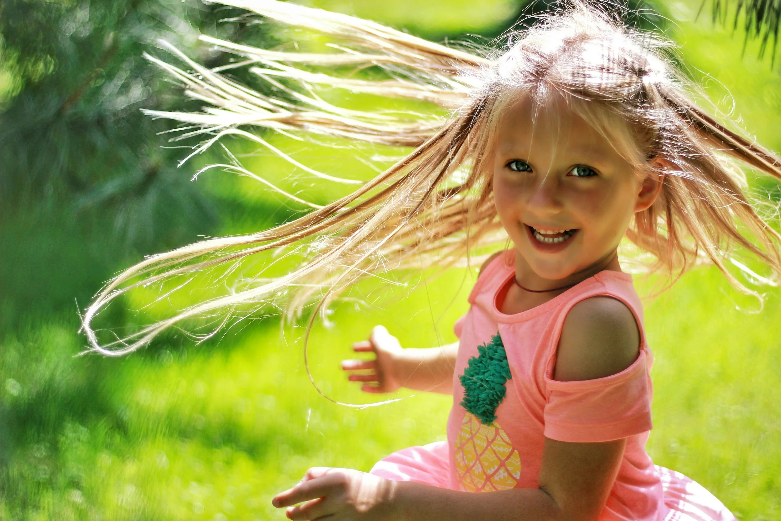 A young girl with blonde hair and blue eyes smiling and looking at the camera while playing outside on a sunny day. She is wearing a pink sleeveless dress with a pineapple design and green ruffle details, surrounded by green grass and trees.