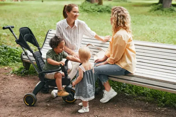 Two women sitting on park benches with three children, one child in a stroller, in a green outdoor park.