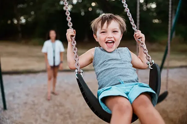 A young boy smiling and enjoying a ride on a swing in a park, with a girl standing in the background.