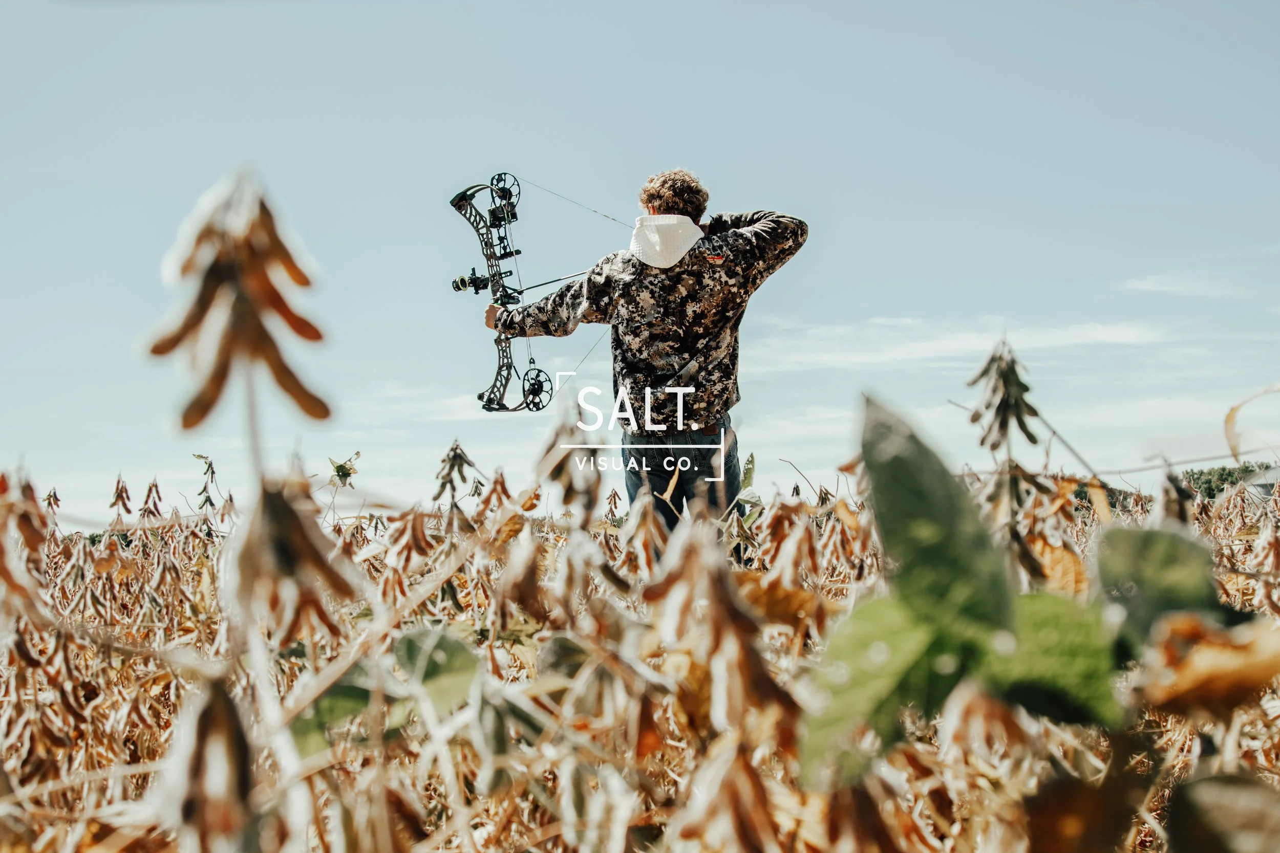 A person with curly hair wearing camouflage clothing and a white hoodie, holding a compound bow, standing in a field of dried corn plants under a clear blue sky.