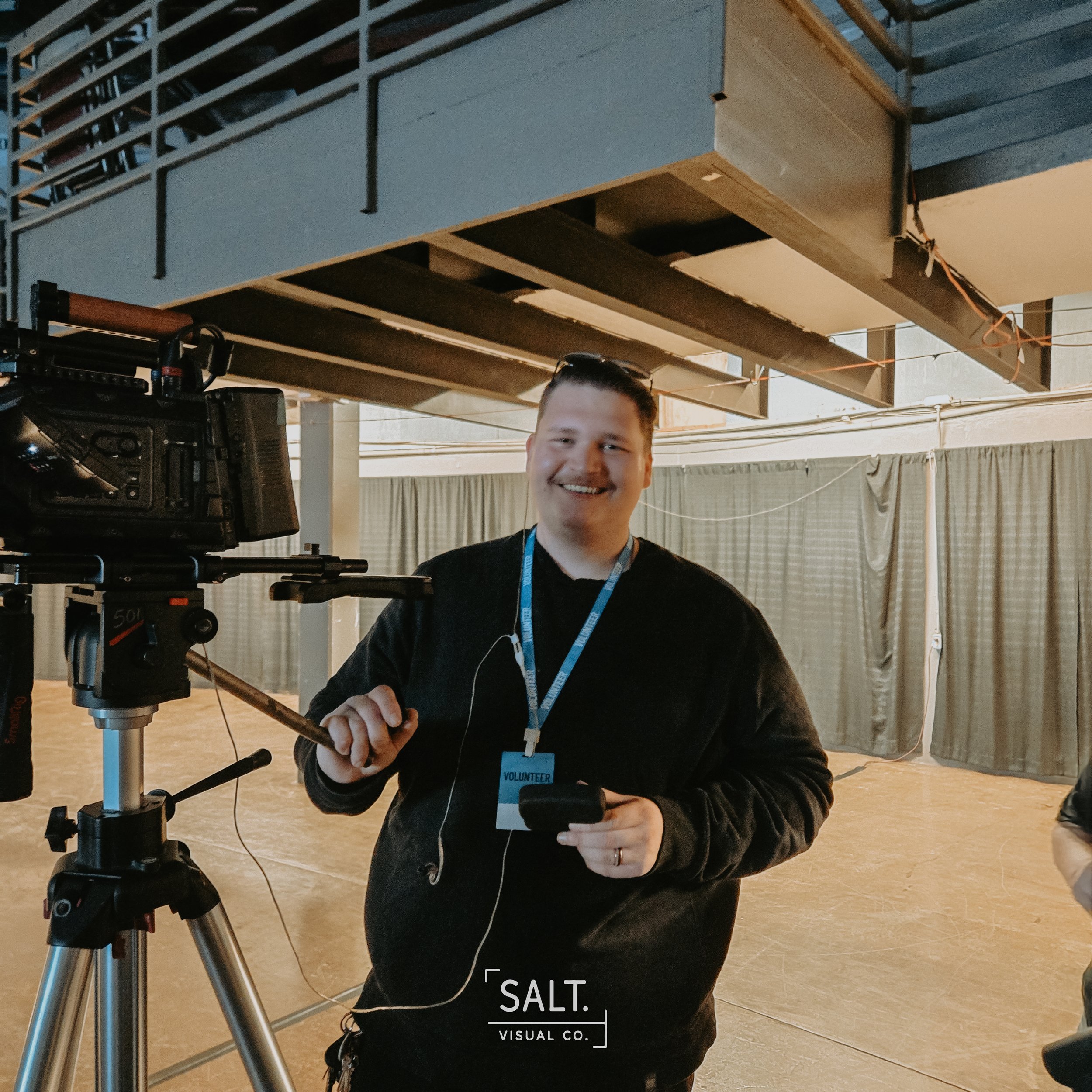 A smiling man wearing a black shirt and a volunteer badge standing next to a camera on a tripod inside a building with scaffolding and curtains.