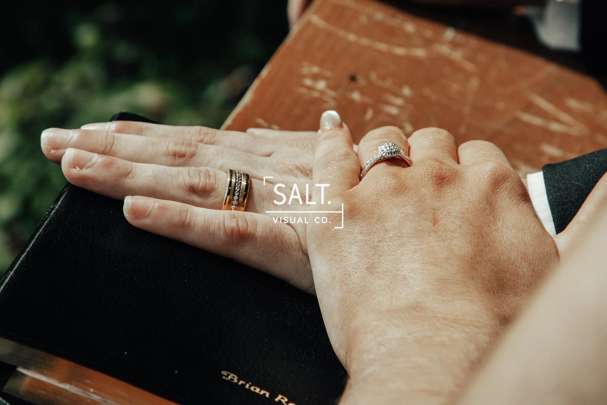 Close-up of two hands with wedding rings resting on a black surface, with a wooden box in the background.
