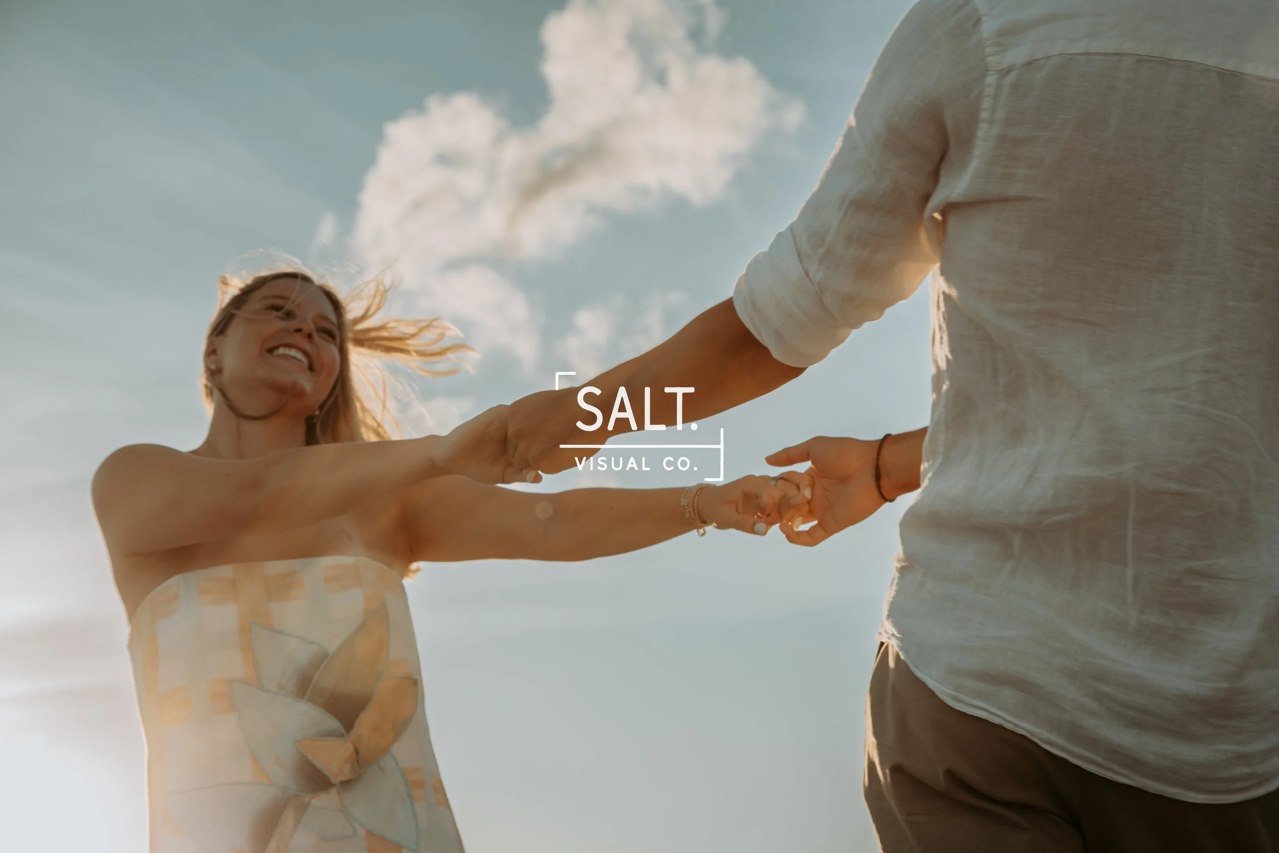 A woman with blonde hair smiling and holding hands with a man outside, both dressed in light, summery clothing, with the sky and clouds in the background, during daylight.