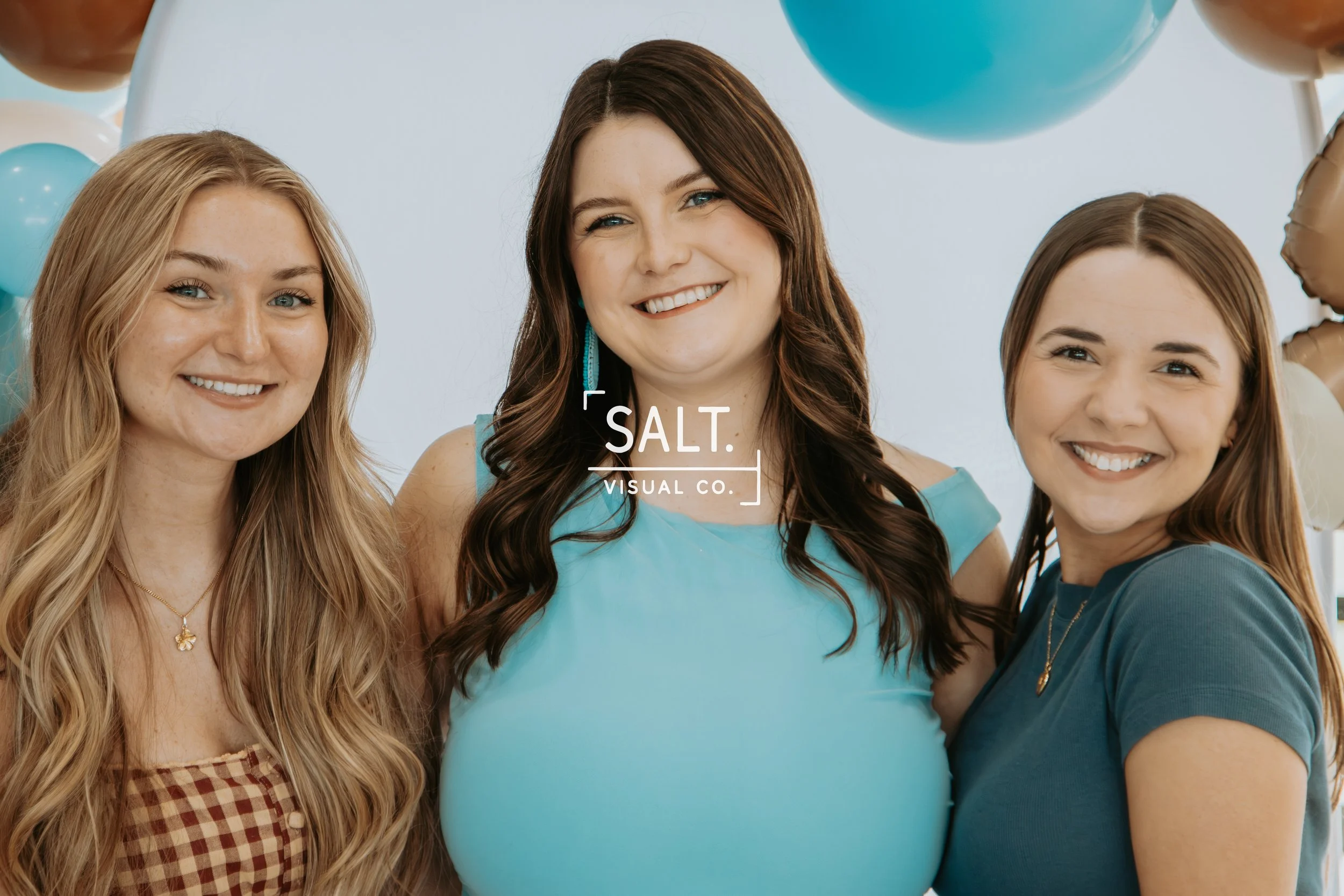 Three women smiling, standing close together, with balloons in the background. The woman in the center has dark brown hair and is wearing a blue top. The woman on the left has long, wavy blonde hair and is wearing a checkered shirt. The woman on the right has brown hair and is wearing a dark top.