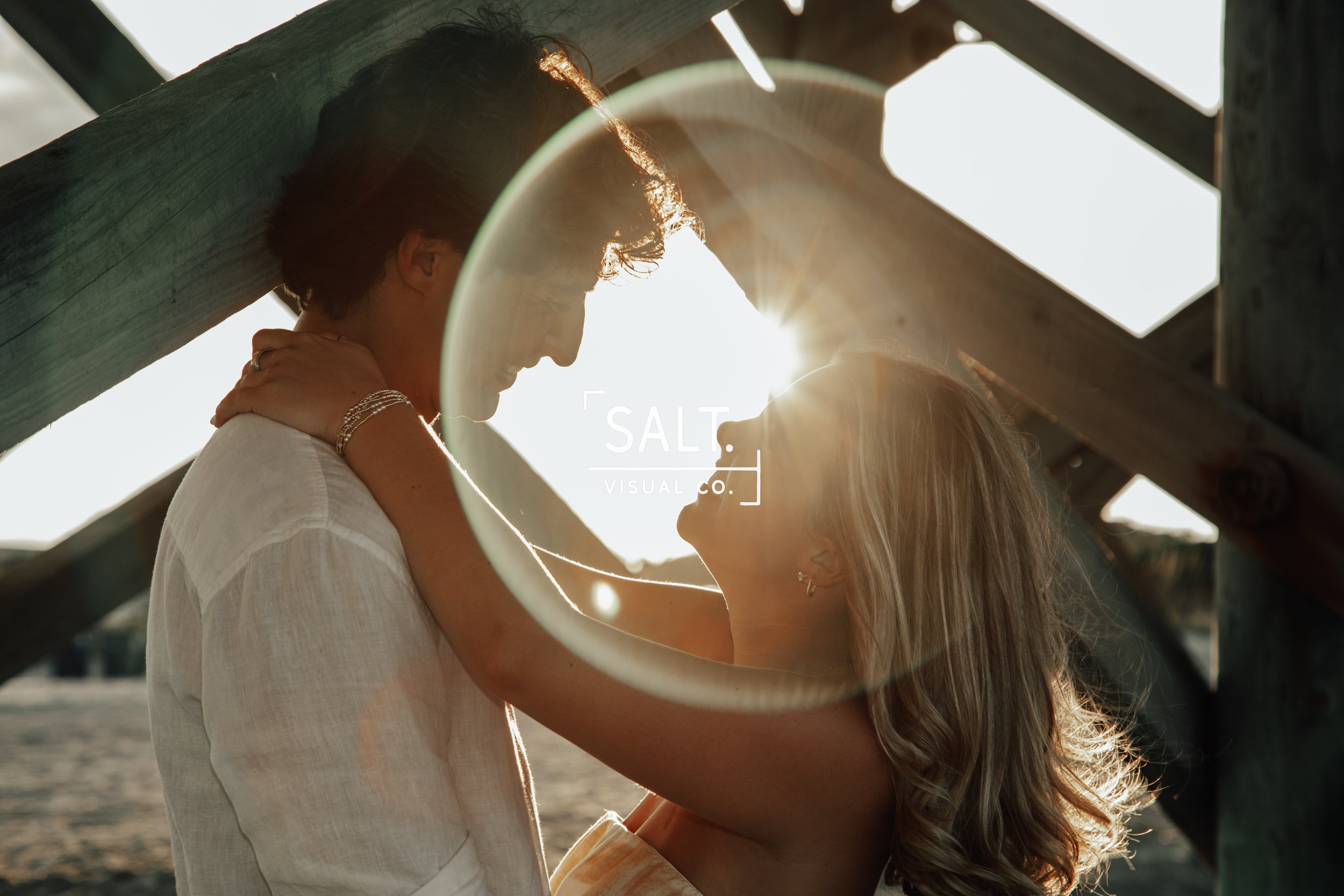 A couple stands close together outdoors beneath a wooden structure, with sunlight shining between them, creating a backlit scene. The man has dark hair and the woman has long, wavy blonde hair, and they are looking into each other's eyes.