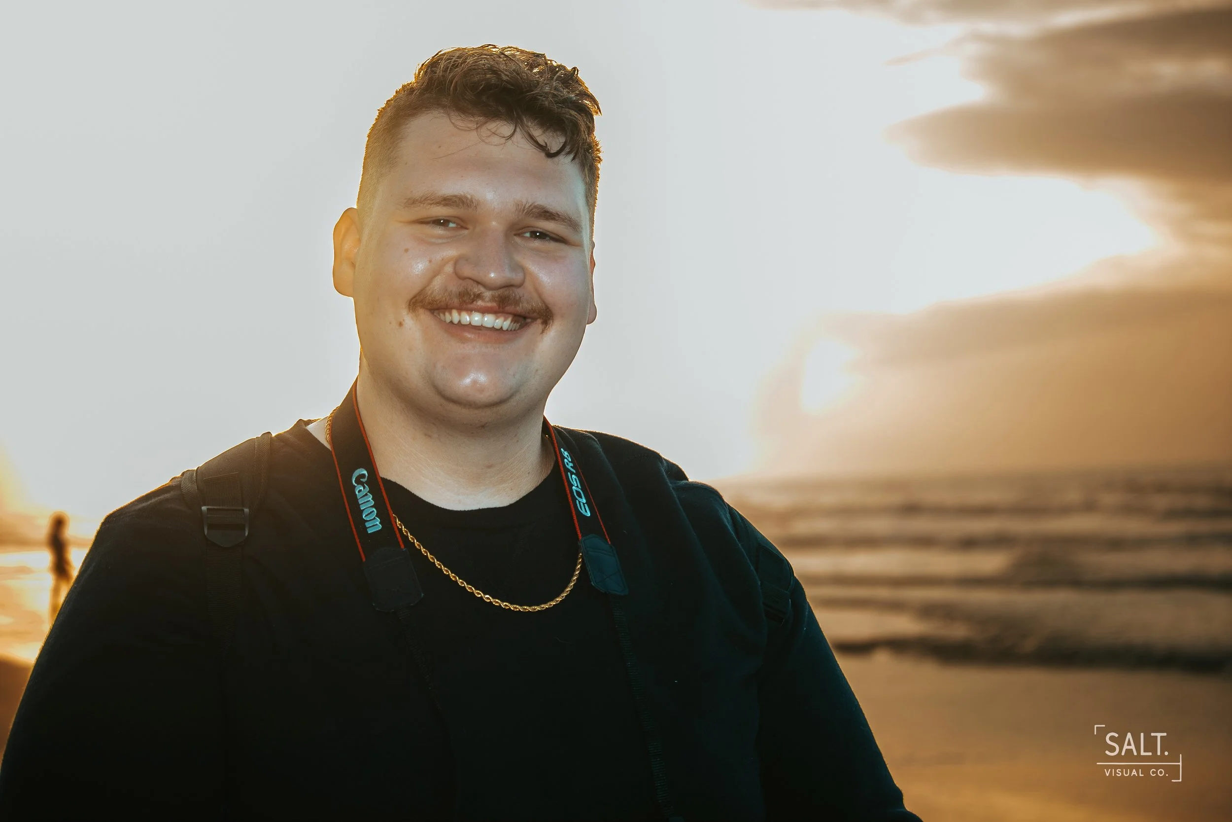 A young man smiling at the camera at sunset by the water, wearing a black shirt, a gold chain, and a camera around his neck.