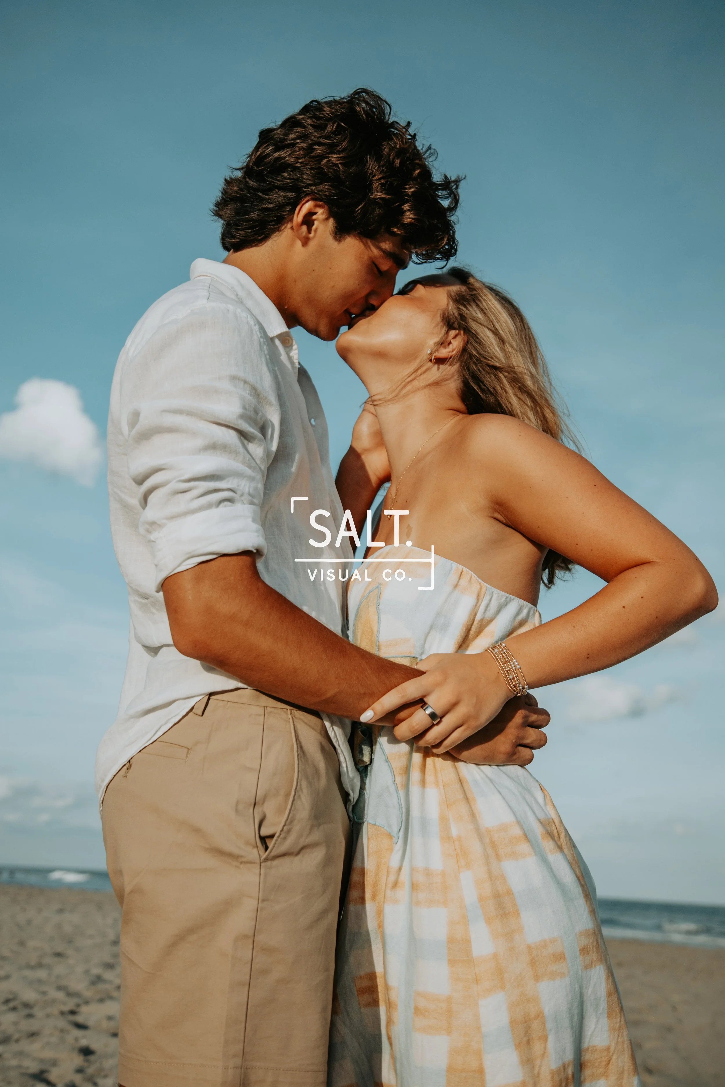 A young couple kissing on the beach during the daytime, with the man wearing a white shirt and beige shorts, and the woman in a light-colored sundress, sunlight illuminating them, blue sky and clouds in the background.