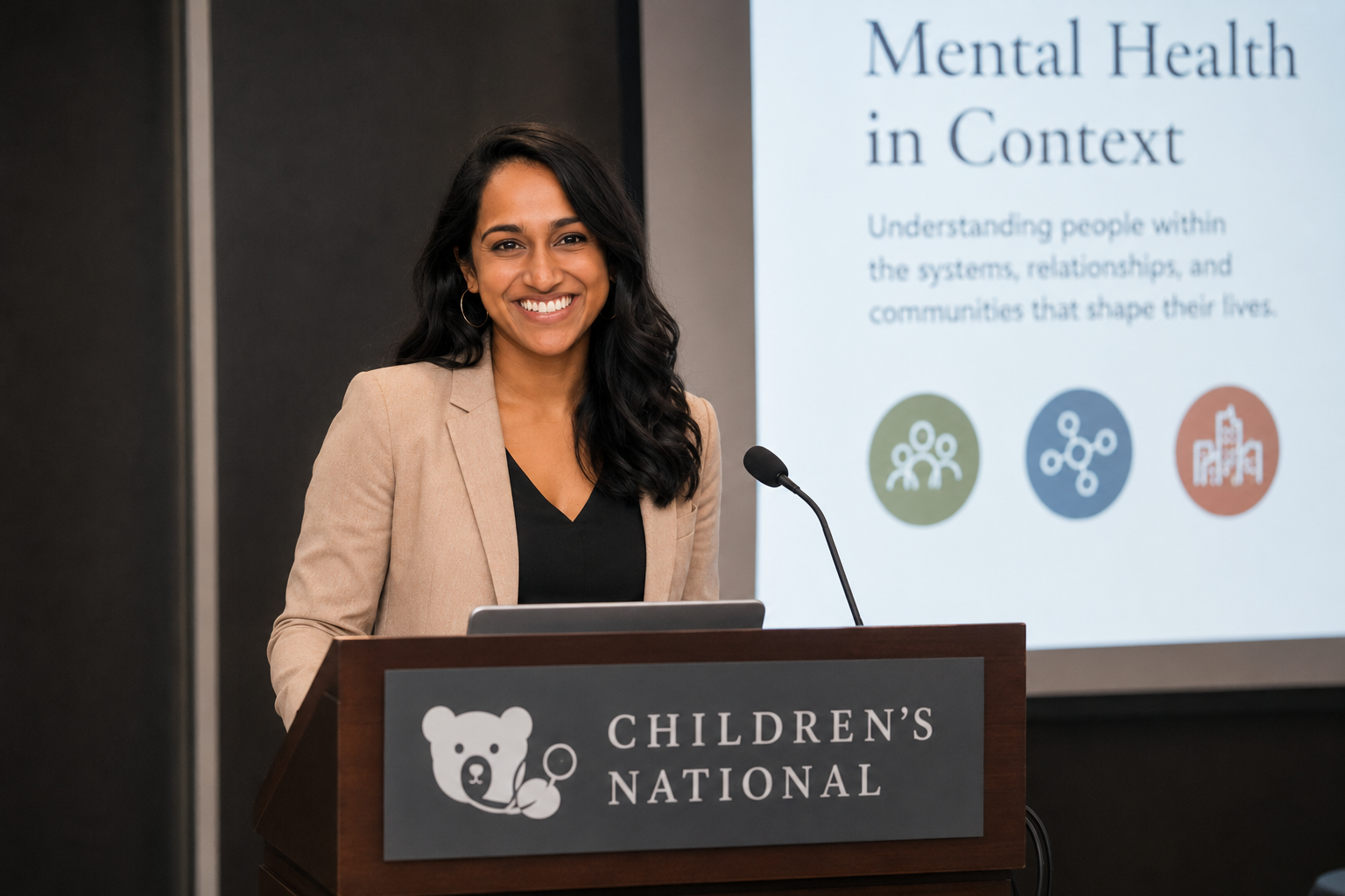 A woman standing at a podium labeled 'Children's National' giving a presentation on mental health. She is smiling, wearing a beige blazer and black top, with dark wavy hair. Behind her is a large screen displaying a slide titled 'Mental Health in Context' with text and icons about understanding people within systems, relationships, and communities.