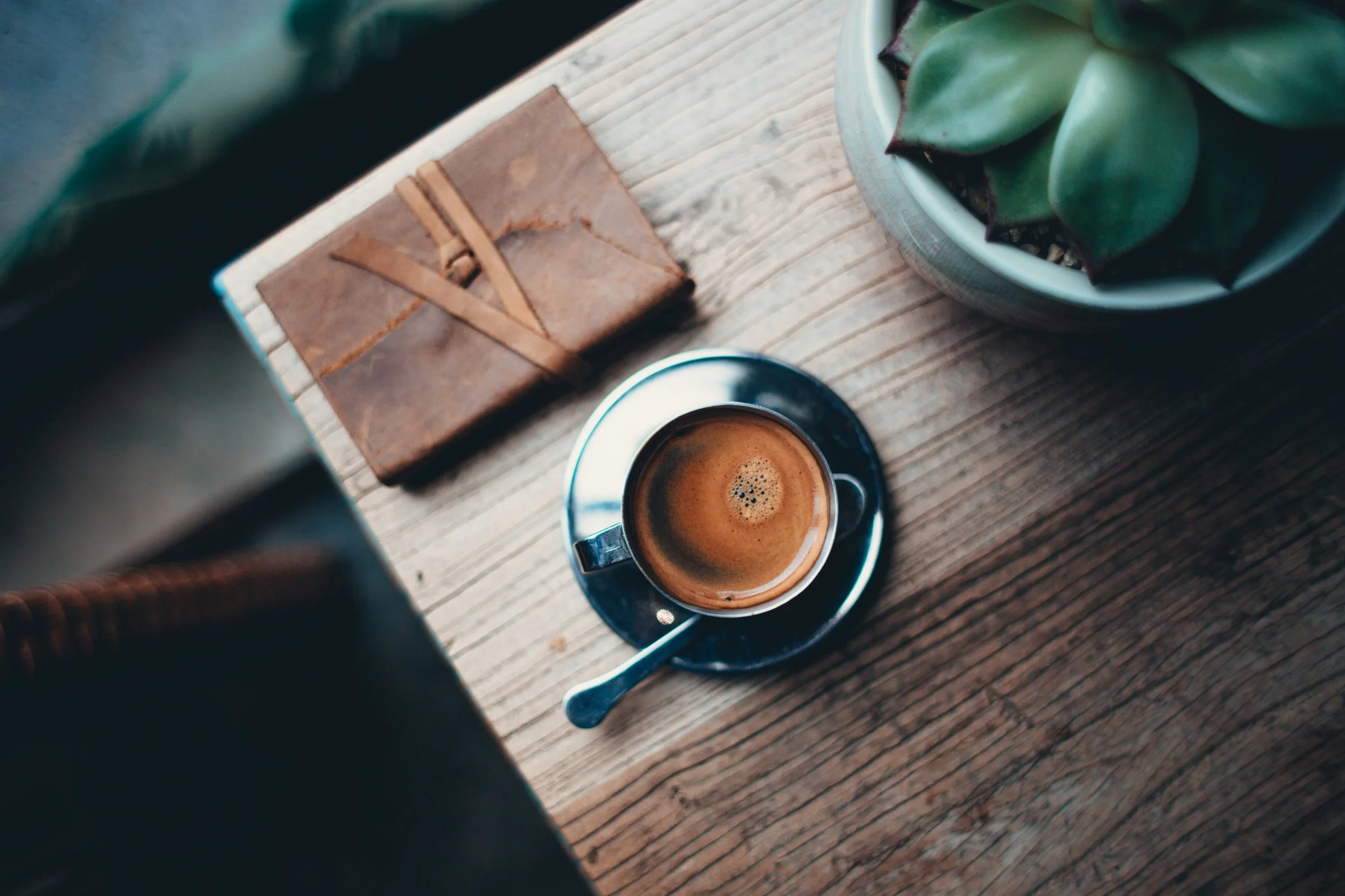A top view of a wooden table with a cup of espresso, a small leather notebook, and a potted succulent plant.