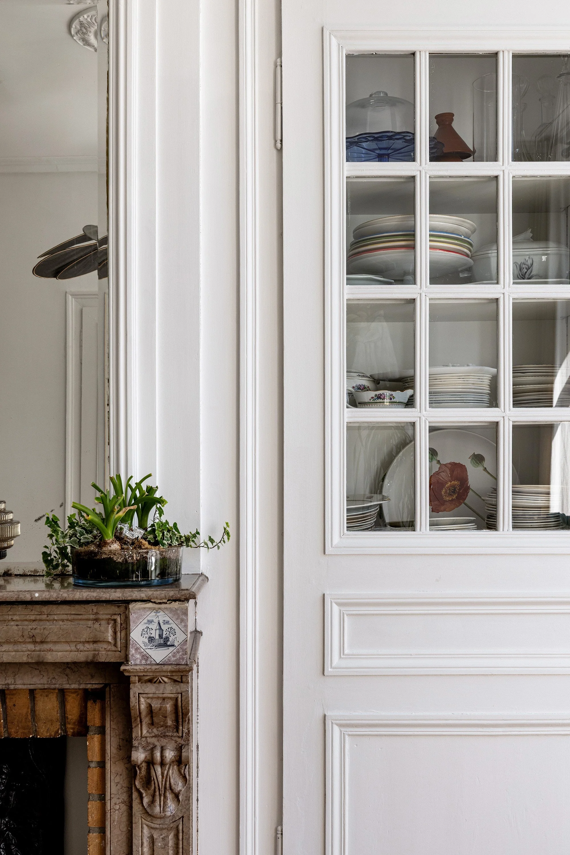 Une étagère en verre contenant des assiettes, des bols et des carafes. À côté, une cheminée ancienne en marbre avec une plante verte en pot placé dessus.