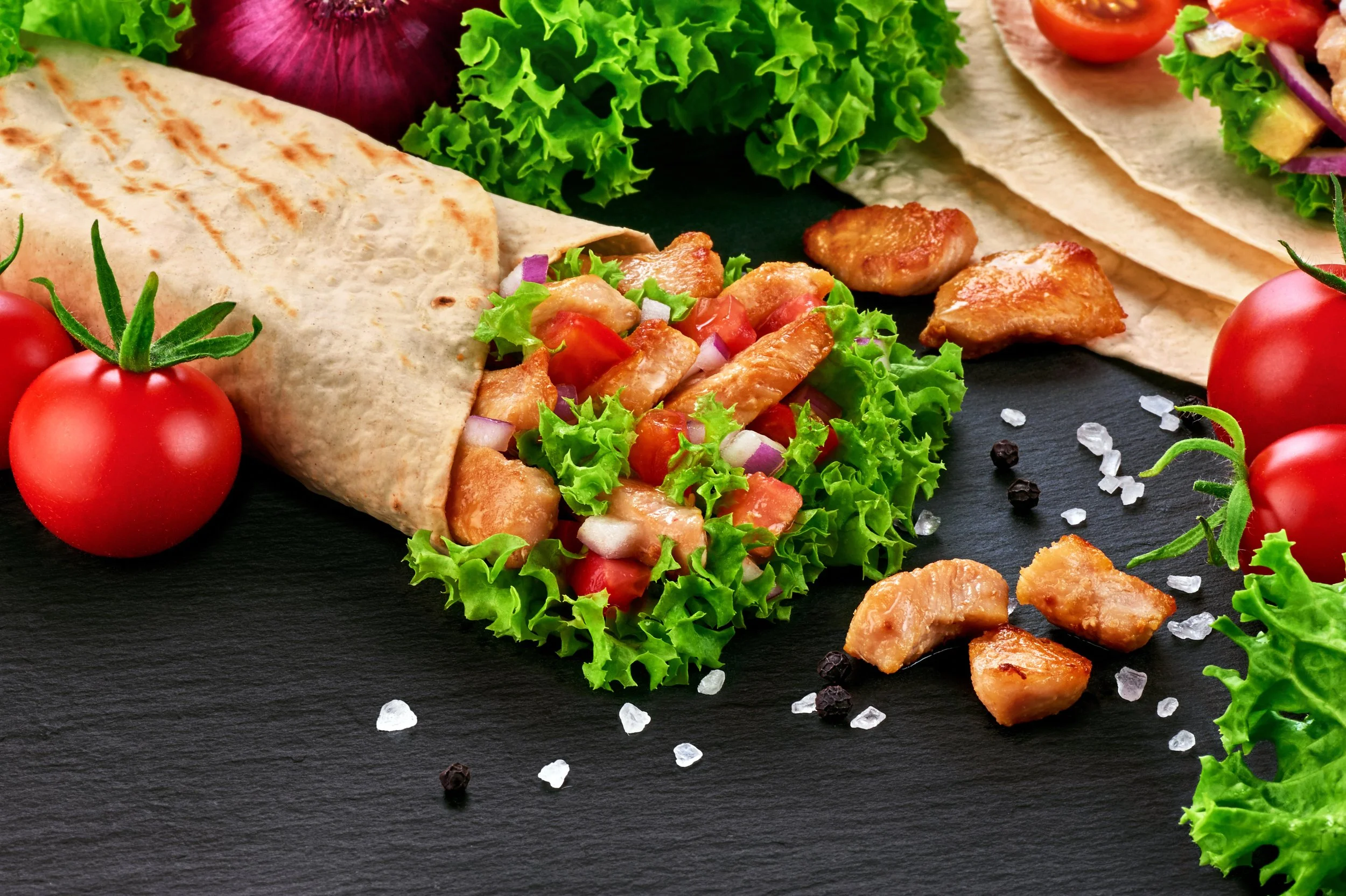 Close-up of a burrito on a dark surface, surrounded by cherry tomatoes, lettuce, avocado, red onion, and salt and pepper grains.