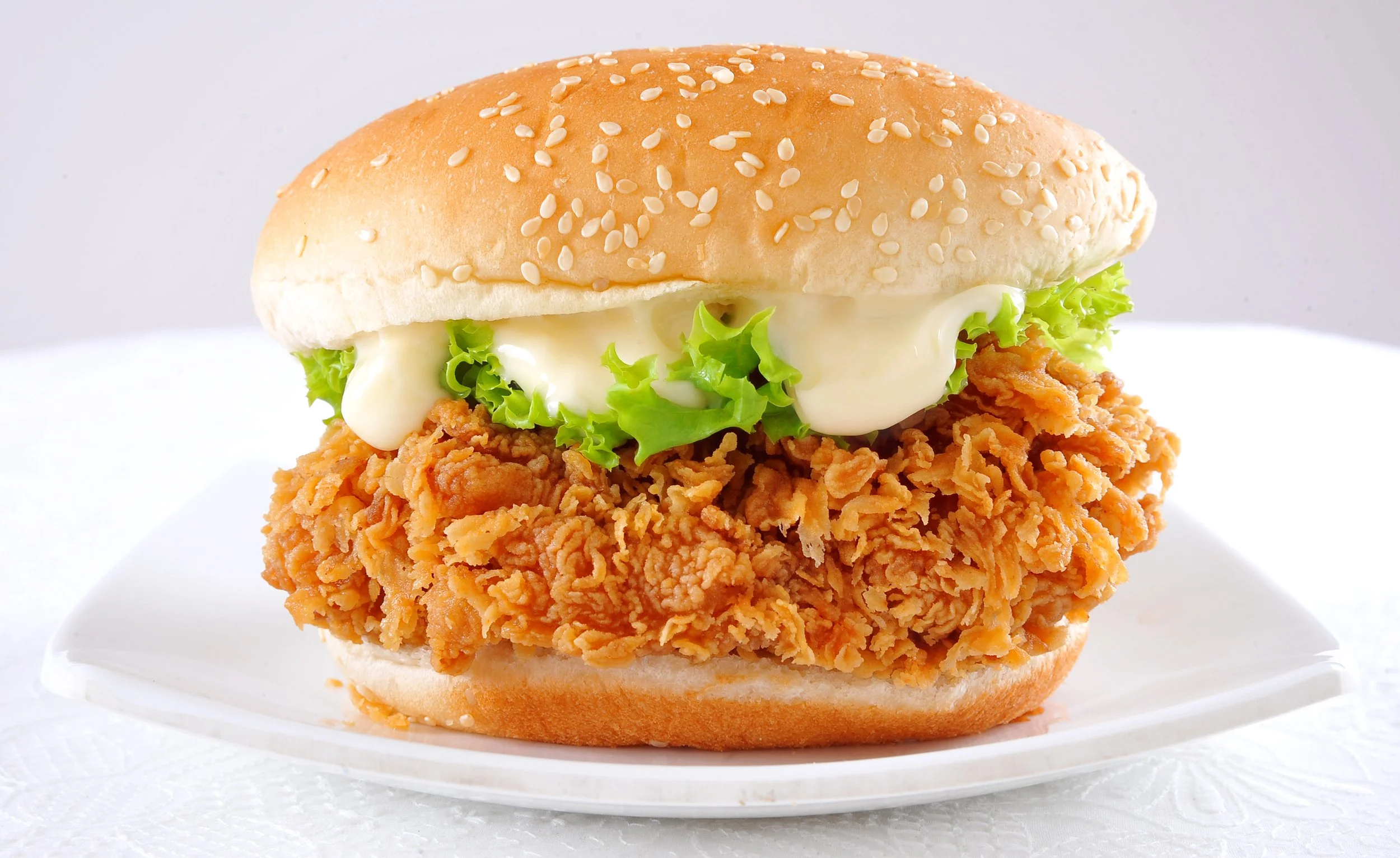 Fried chicken sandwich with lettuce, mayonnaise, and sesame seed bun on a white plate.