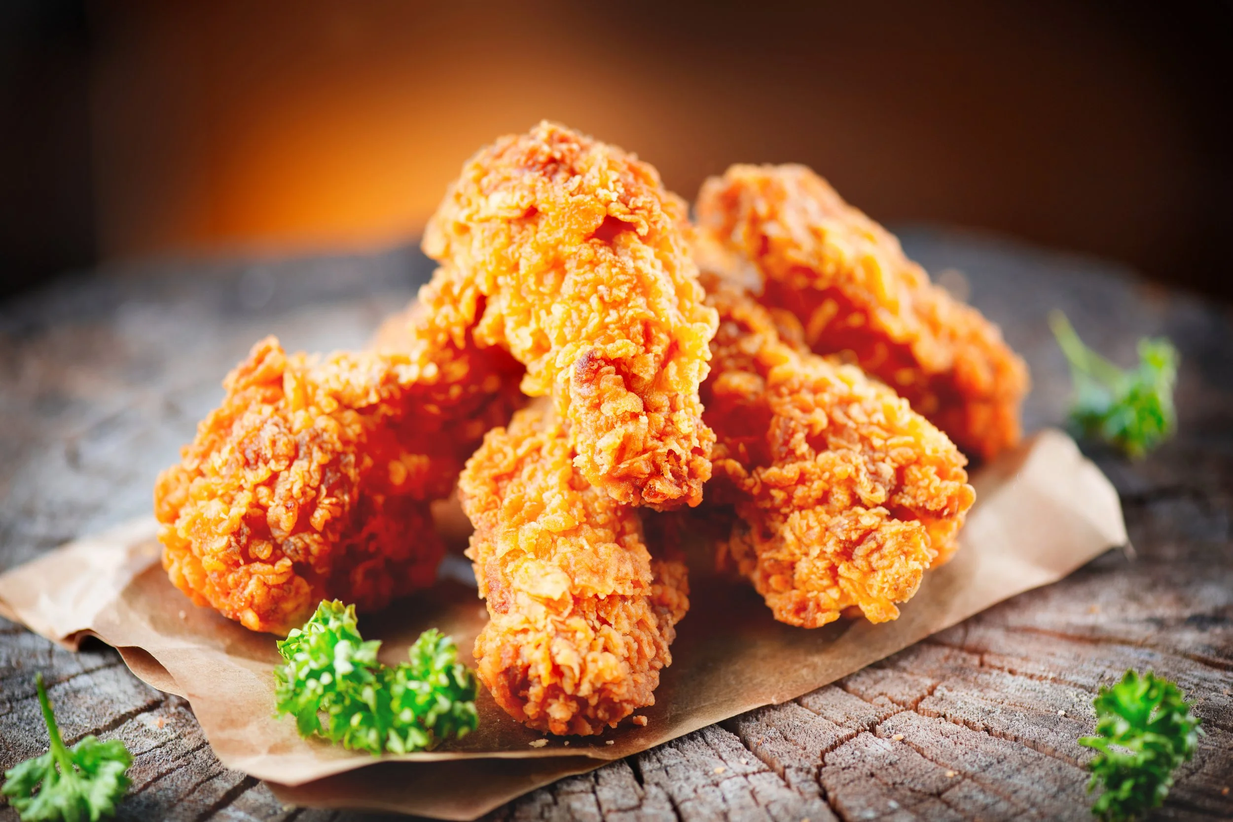 Close-up of crispy fried chicken drumsticks on brown paper on a rustic wooden surface, garnished with parsley.