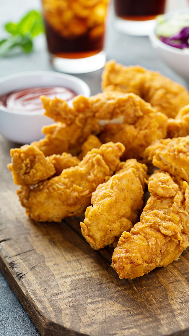 Close-up of fried chicken tenders on a wooden serving board with dipping sauces and glasses of soft drinks in the background.