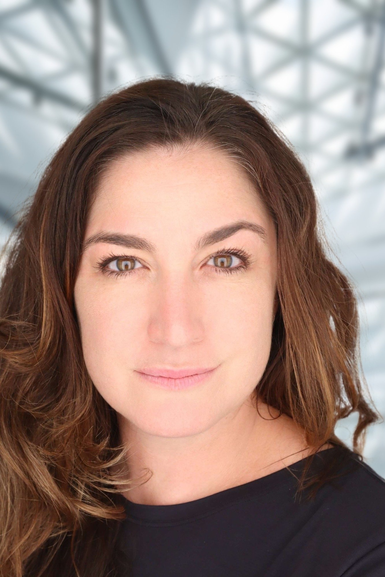 Close-up of a woman's face with brown hair and light skin, wearing a black top, in front of a blurred background with a metallic structure.