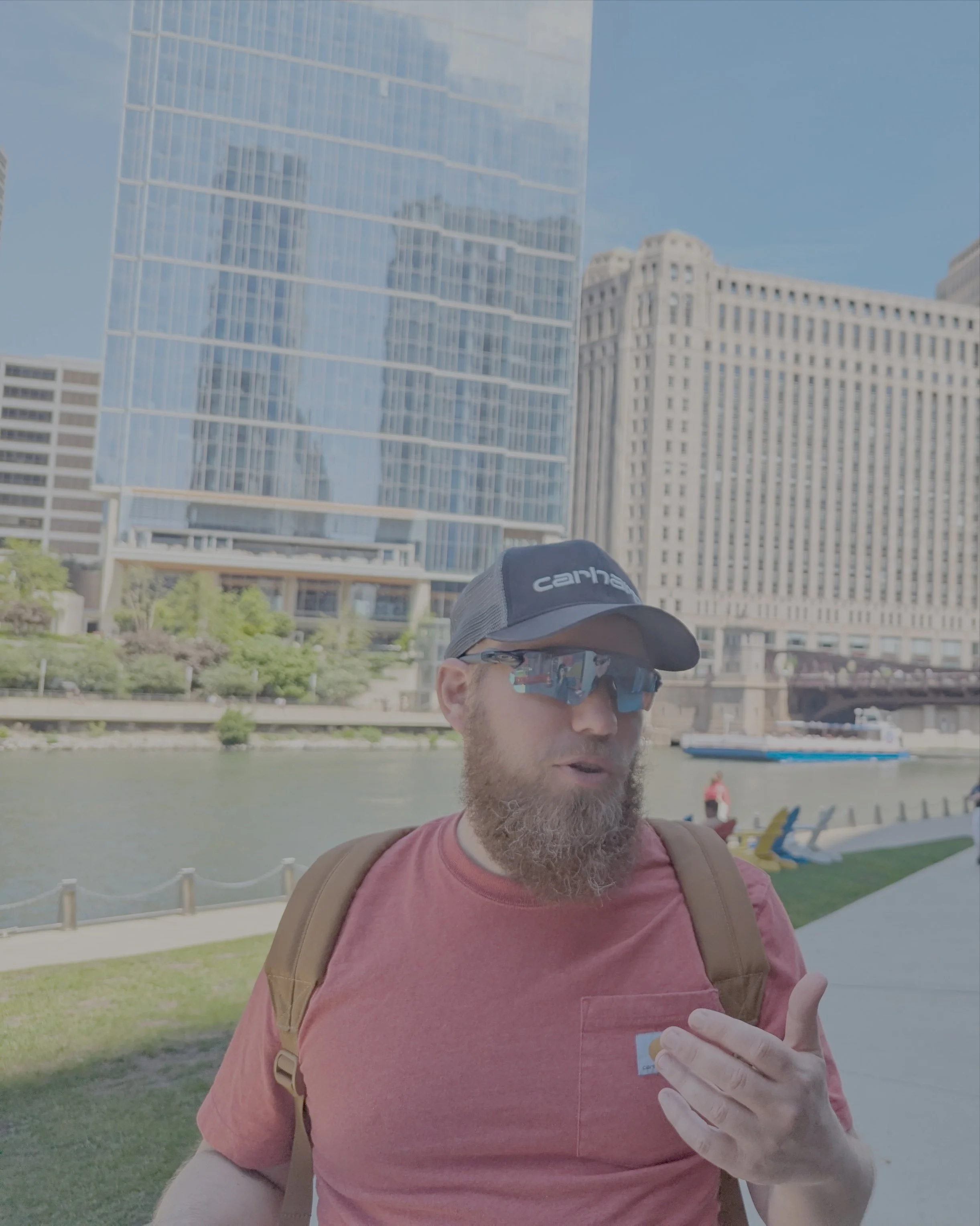 A man with a beard wearing sunglasses, a cap, and a pink t-shirt, standing outdoors in front of a grassy area with trees and a backdrop of tall modern glass buildings.