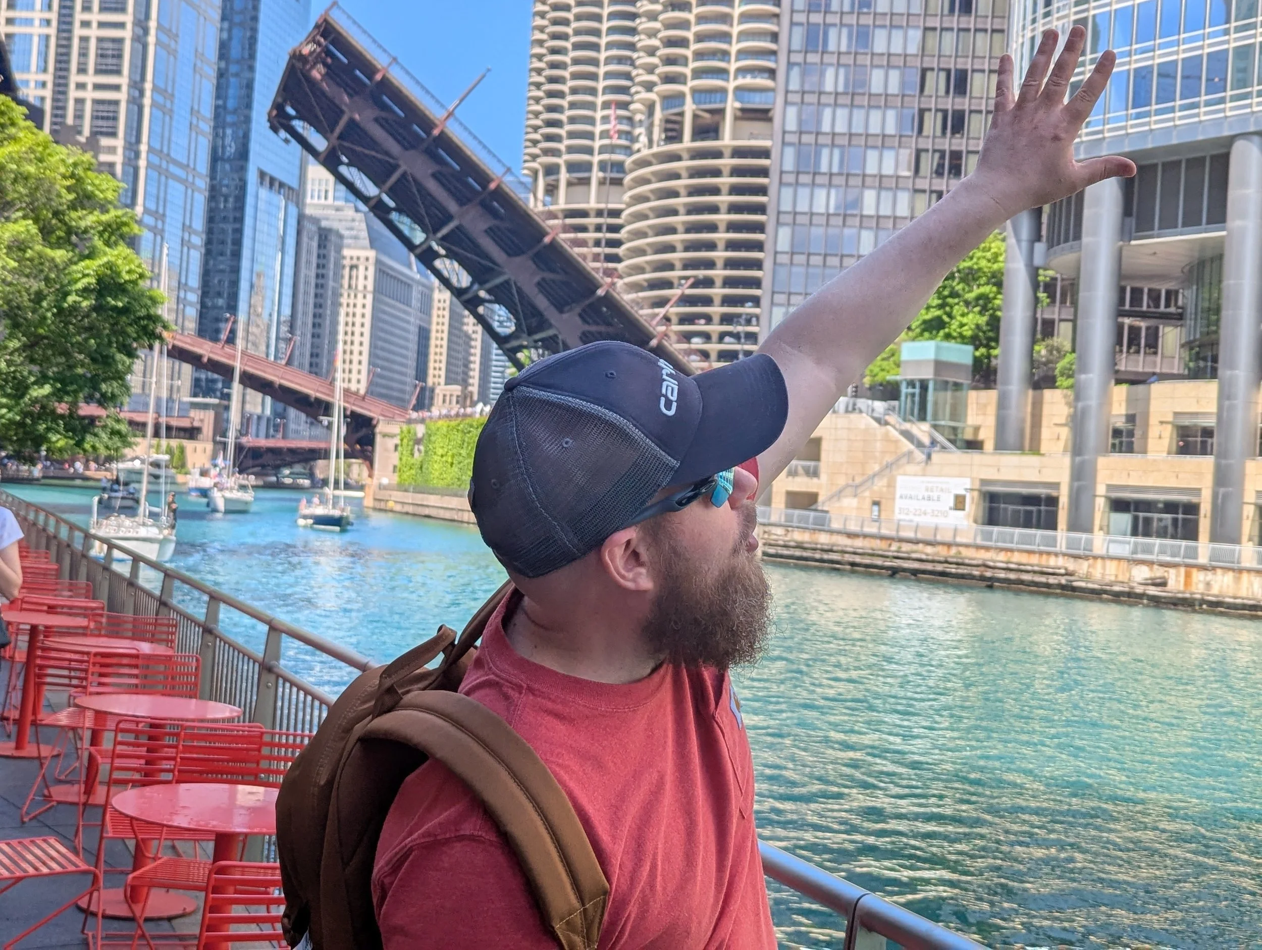 A man with a beard wearing a red shirt and a black cap, carrying a backpack, standing by a waterfront with boats and modern high-rise buildings in the background, reaching out with his right arm.