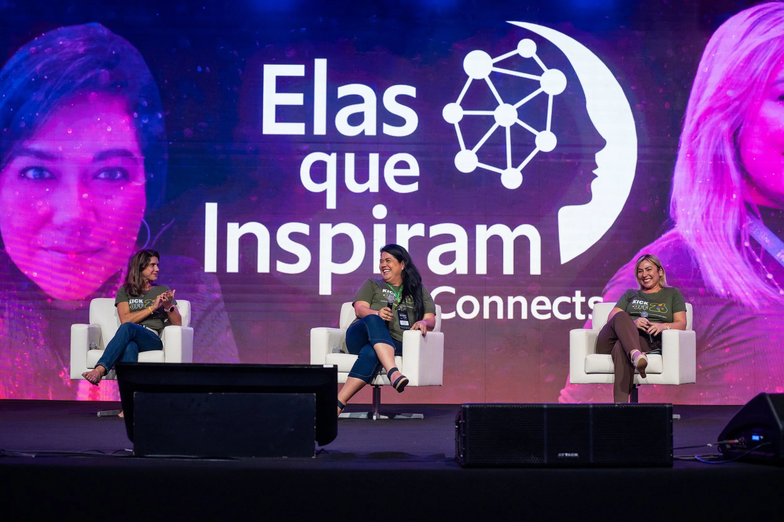 Três mulheres sentadas em cadeiras brancas em um palco de conferência, com fundo digital mostrando o texto "Elas que inspiram, She Connectst" e imagens de retratos femininos.