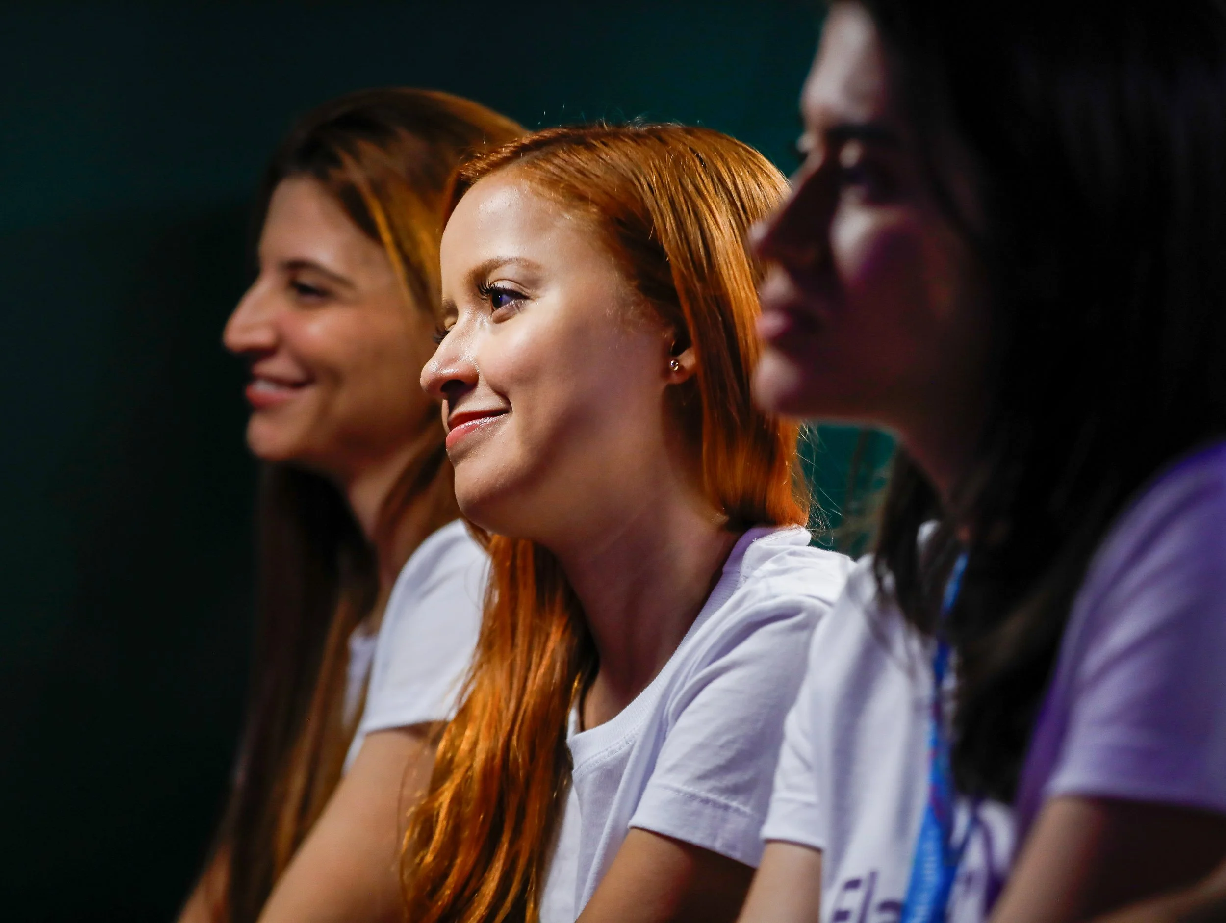 Imagem de três jovens mulheres sorrindo, todas de perfil, com destaque para a jovem de cabelo vermelho no centro, usando uniforme branco.