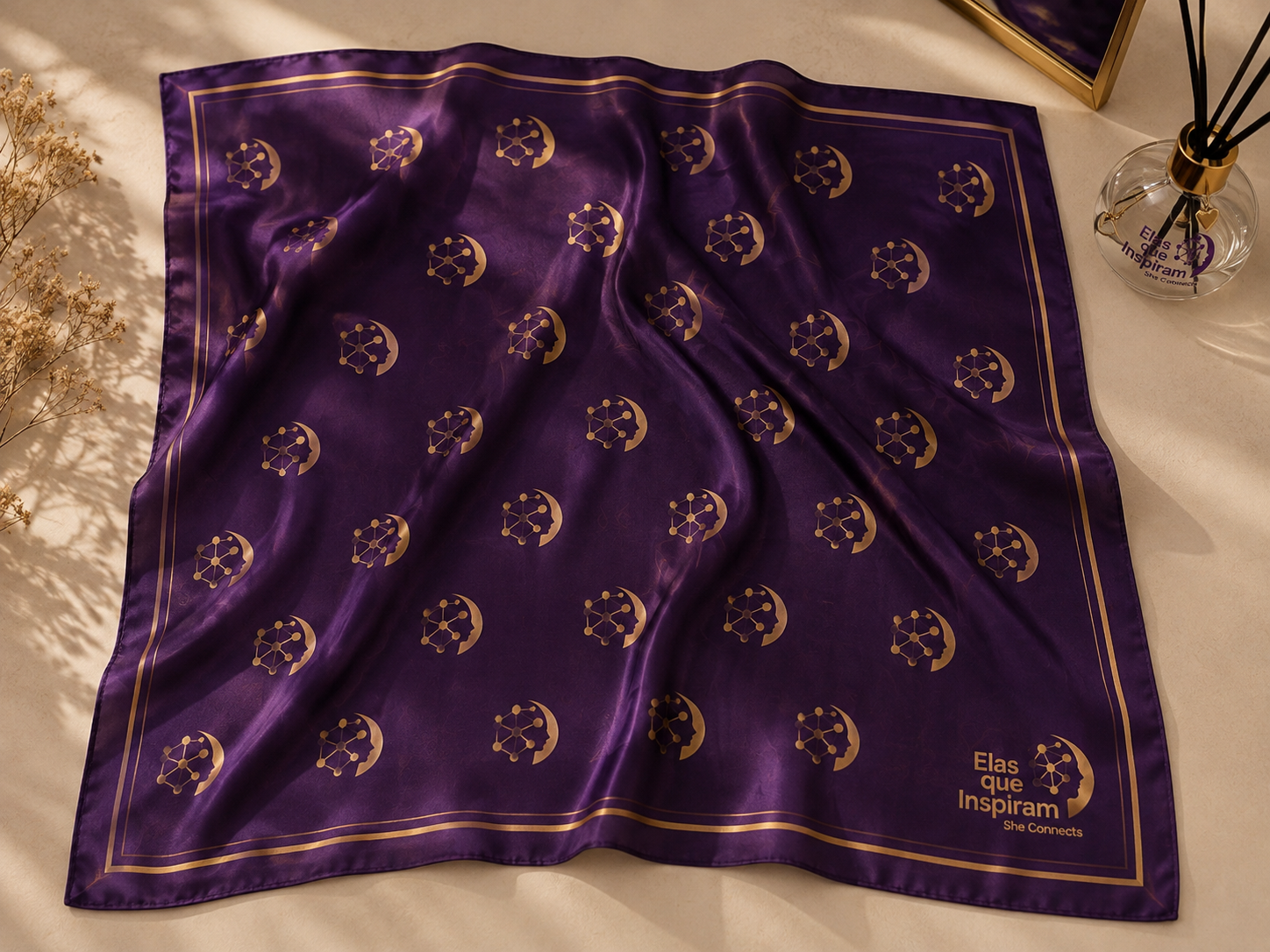 Lenço roxo com padrão de símbolos de constelações e luas em dourado, com uma borda dourada fina. Ao lado, um vaso com óleo essencial e ramos secos, e uma moldura de espelho dourada.