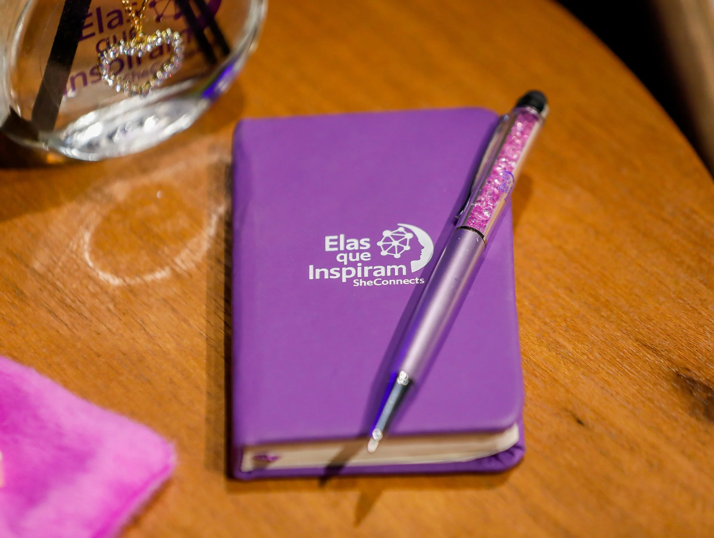 Imagem de uma mesa de madeira com um livro roxo intitulado 'Elas que Inspiram', uma caneta de ponta preta com detalhes rosa brilhante, uma taça de vidro parcialmente visível com um colar de pérolas dentro e uma parte de uma mão com pele clara e cabelo rosa na margem inferior da imagem.