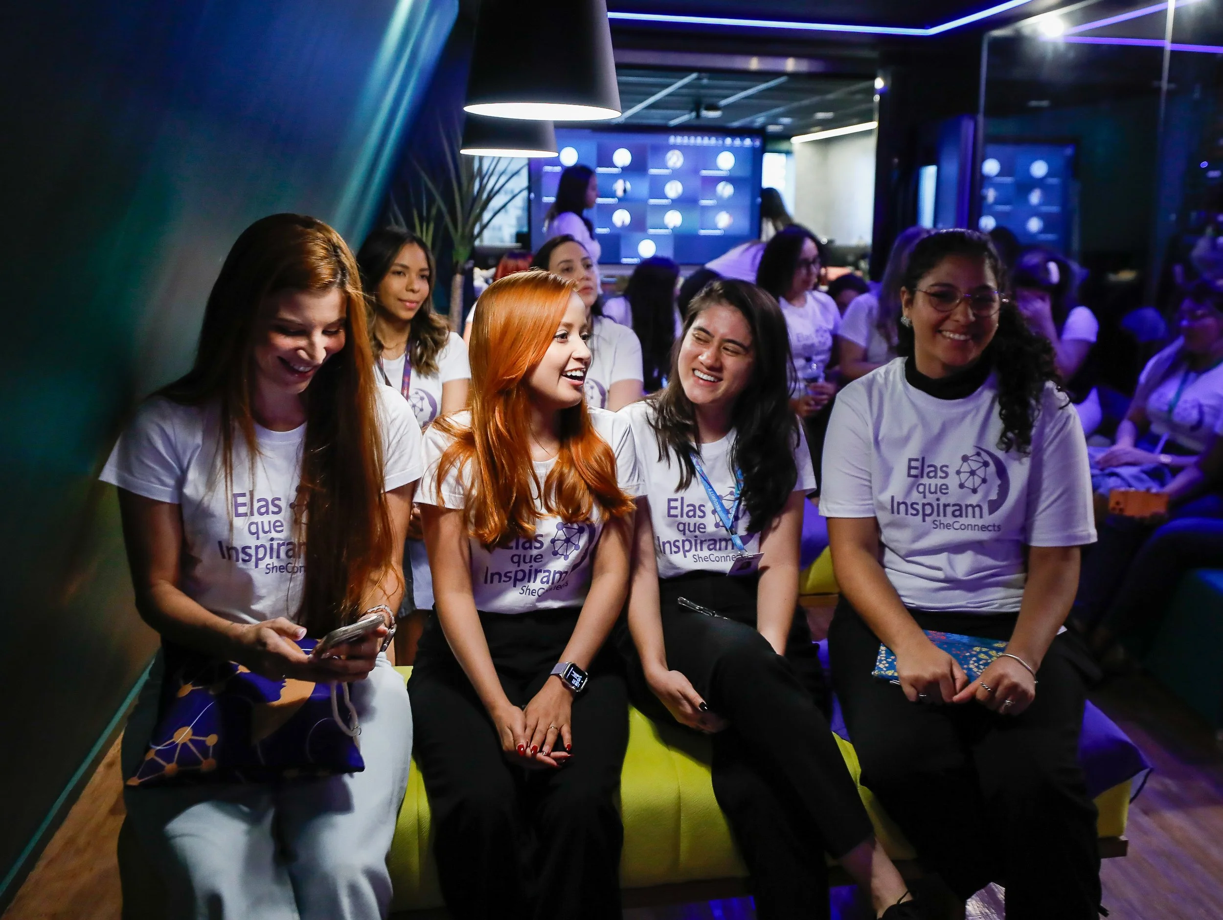 Grupo de mulheres sorrindo e conversando em um ambiente de evento ou conferência com iluminação azul e fundo com telas digitais.