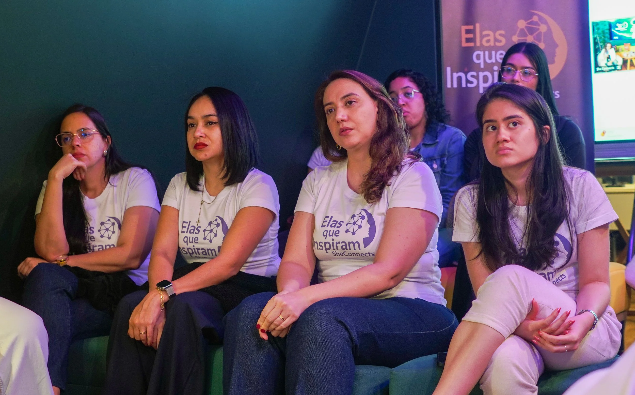 Grupo de quatro mulheres sentadas assistindo uma apresentação, vestindo camisetas brancas com o texto 'Elas que Inspiram' e logotipo da SheConects, em ambiente com paredes escuras e telas de fundo.